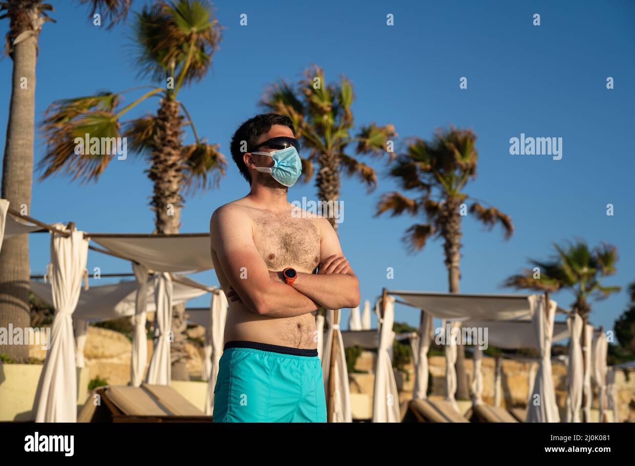 Ruhender Mann in einer Schutzmaske auf einem schmutzigen Strand allein vor dem Hintergrund von Palmen und Sonnenliegen auf dem Mittelmeer Stockfoto