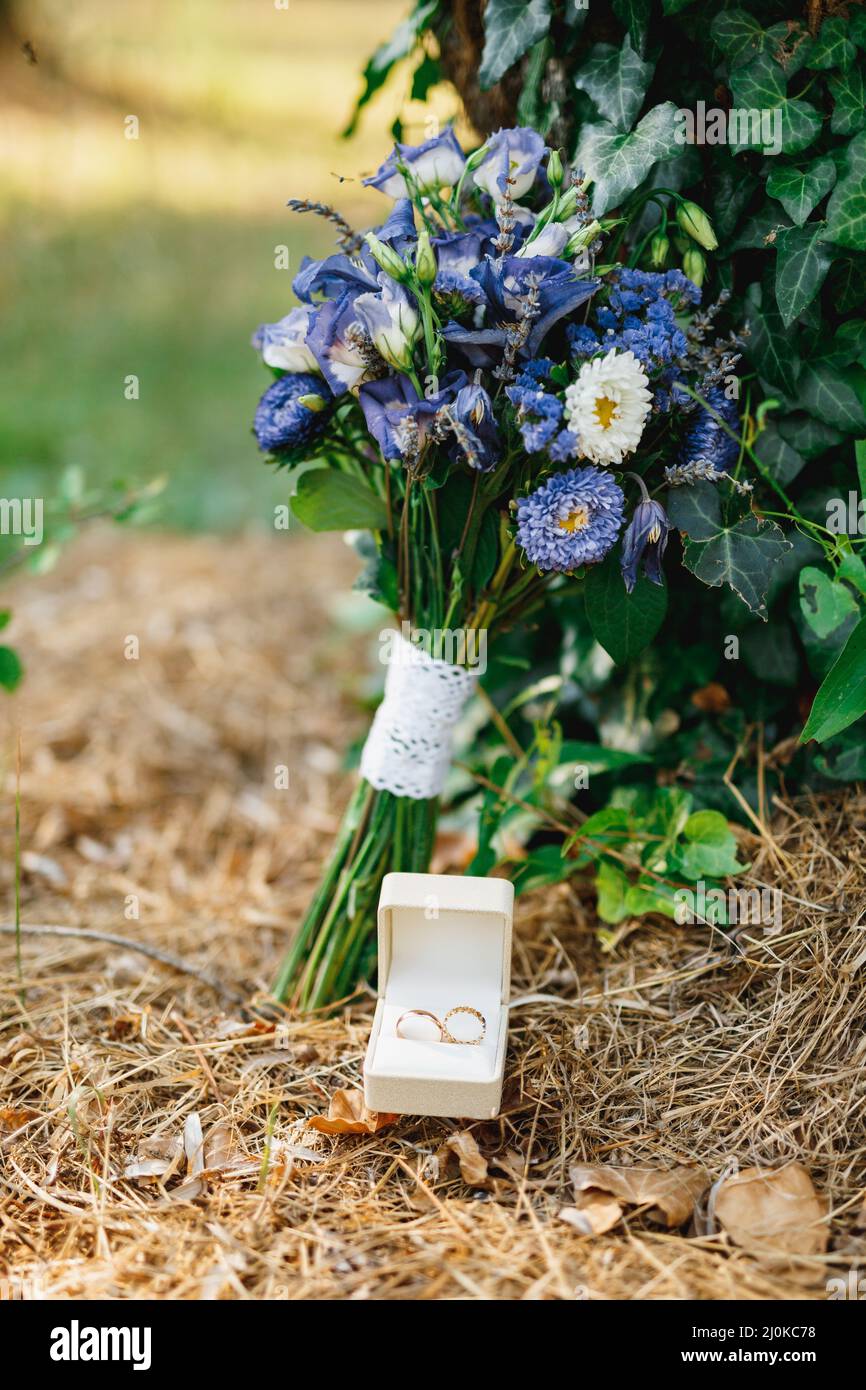 Eheringe in einer weißen Schachtel auf trockenem Gras mit dem Brautstrauß einer Braut aus blauen Blumen. Stockfoto