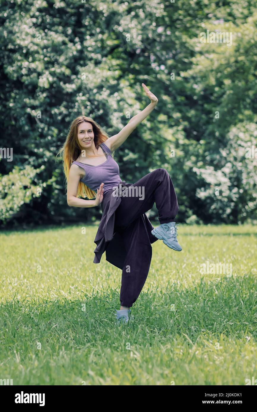 Lächelndes Mädchen im fliederfarbenen Hip-Hop-Outfit in einer Tanzpose auf dem Rasen im Park Stockfoto