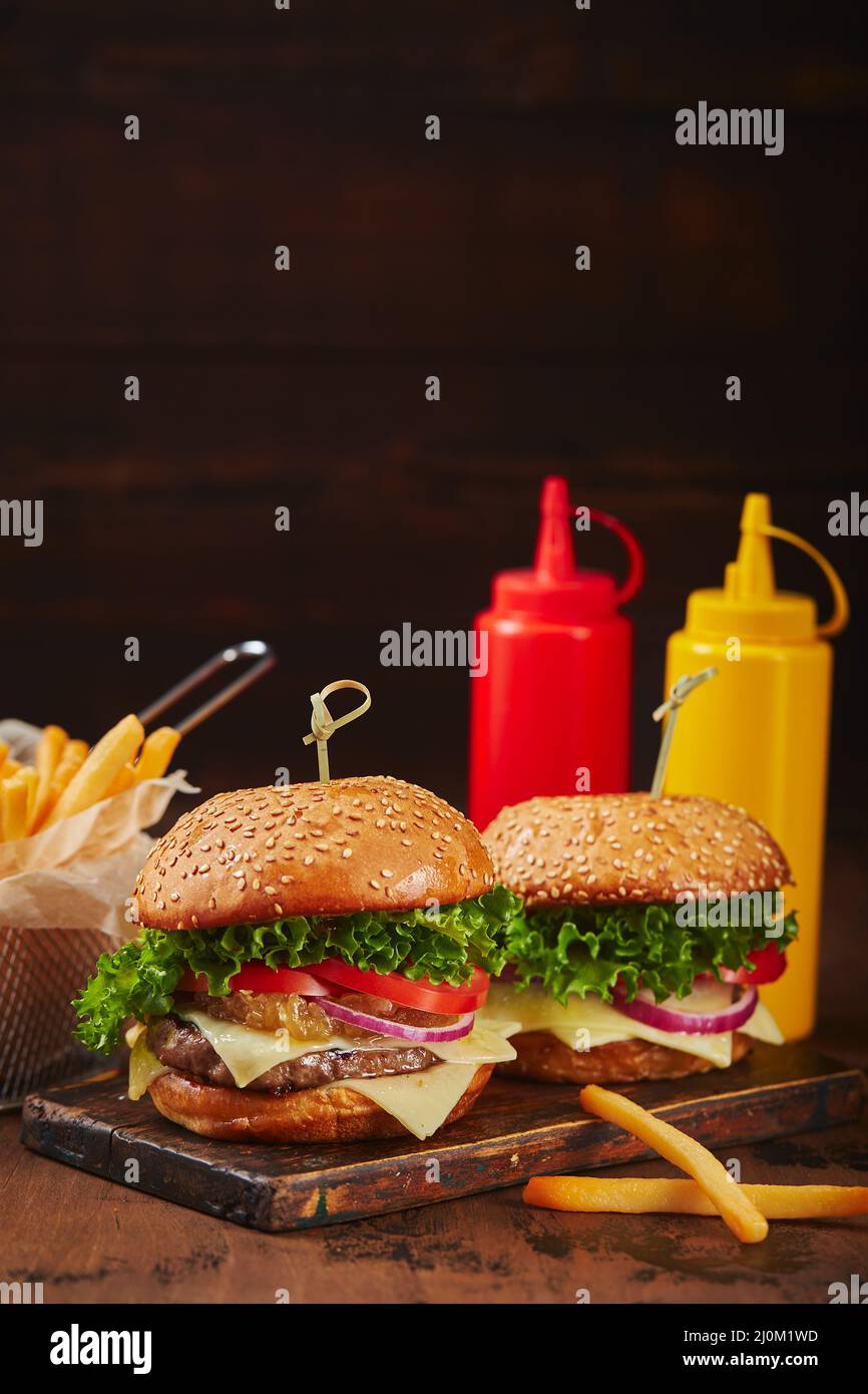 Zwei hausgemachte Burger mit Rind-, Käse- und Zwiebelmarmelade auf einem Holzbrett, Pommes in einem Metallkorb und Saucen. Fast Food Konz Stockfoto