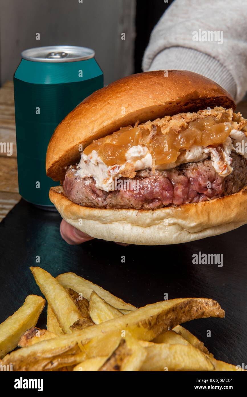 Eine Hand, die ein Stück Rindfleisch-Burger mit viel Frischkäse und karamellisierten Zwiebeln neben einem Stapel hausgemachter Pommes frites hält Stockfoto