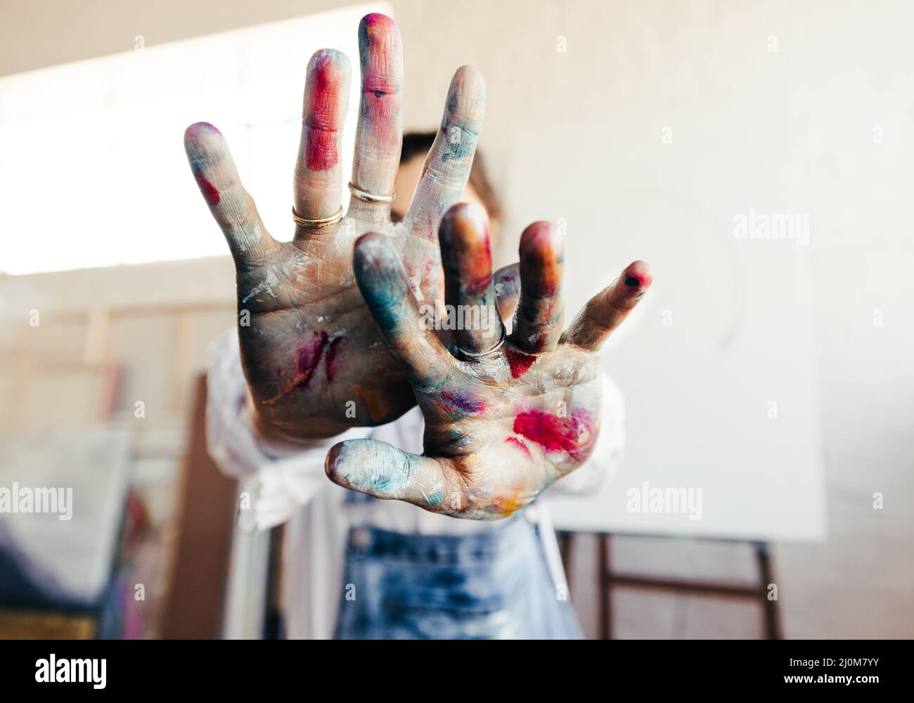 Malerin blockiert die Kamera mit ihren farbigen Händen aus ihrem Gesicht. Nicht erkennbare Künstlerin, die mit ihrer leeren Leinwand in ihrem Atelier steht Stockfoto