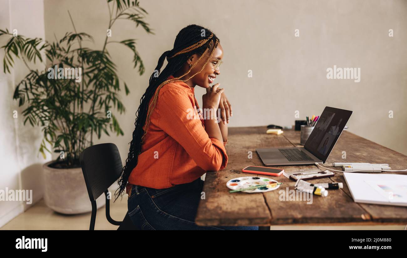 Lächelnde Grafikdesignerin, die an einem virtuellen Meeting teilnimmt, während sie an ihrem Schreibtisch sitzt. Kreative junge Frau, die mit einem Laptop Videoanruf bei ihren Kunden führt. Weiblich Stockfoto