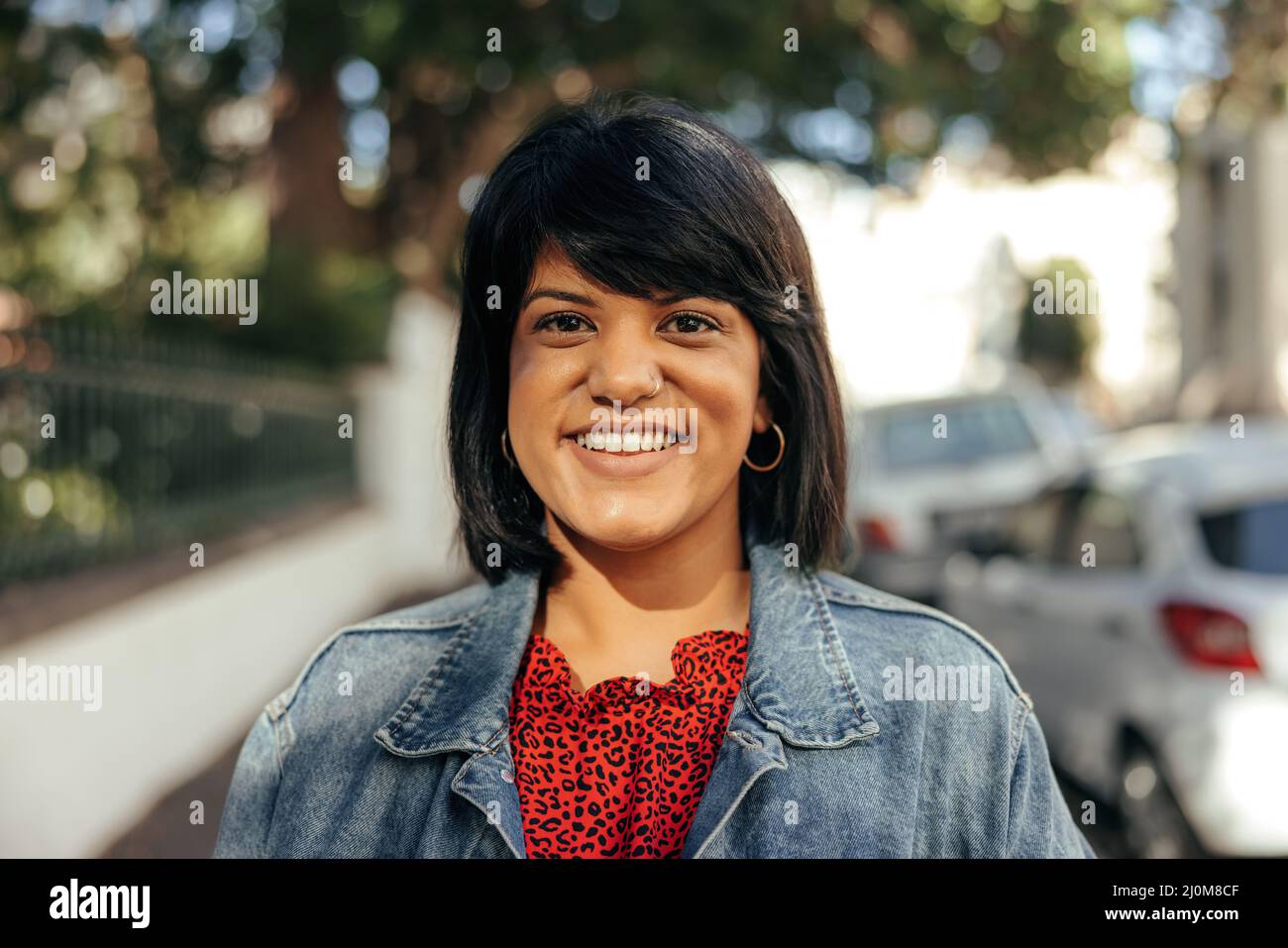 Eine fröhliche Frau mittleren Erwachsenen, die lächelt an der Kamera, während sie auf einem Bürgersteig in der Stadt steht. Glückliche Frau, die allein neben einem Park im Freien steht. Selbstbewusst Stockfoto