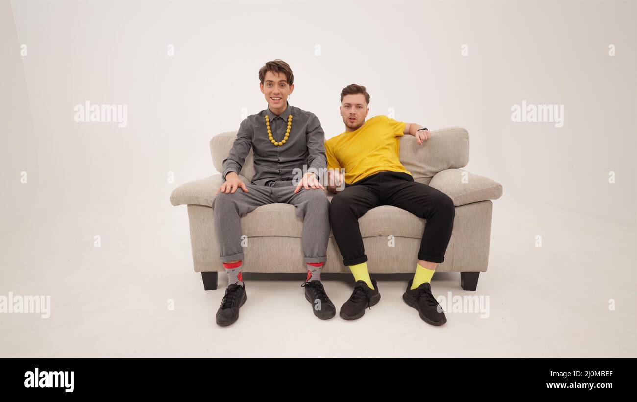 Sehen Sie sich einen Film oder Sportkanal an zwei trendige, hübsche Jungs, die auf einem weißen Sofa auf weißem Hintergrund sitzen. Jungs reagiert auf Stockfoto