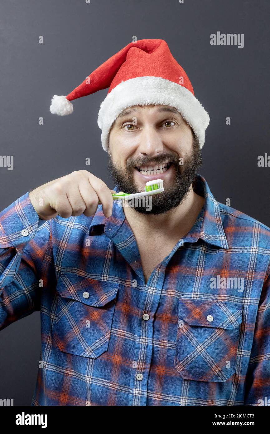 Ein fröhlicher bärtiger Mann in einem Weihnachtsmütze mit einer Zahnbürste. Gefälschter weihnachtsmann. Vorbereitung auf Weihnachten Stockfoto