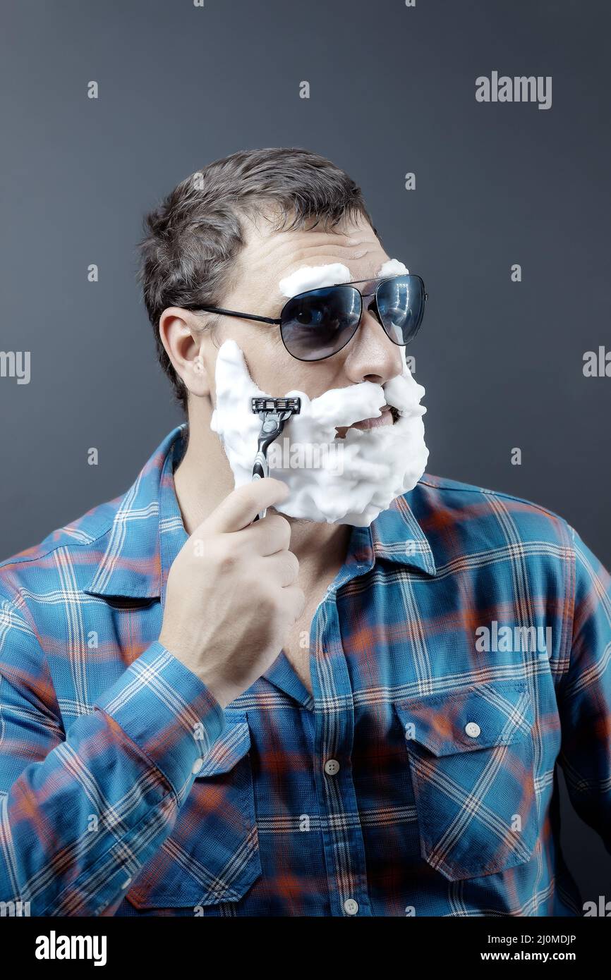 Ein Mann in Sonnenbrille mit Bart und Augenbrauen aus Schaumstoff rasiert mit einem Rasiermesser Stockfoto