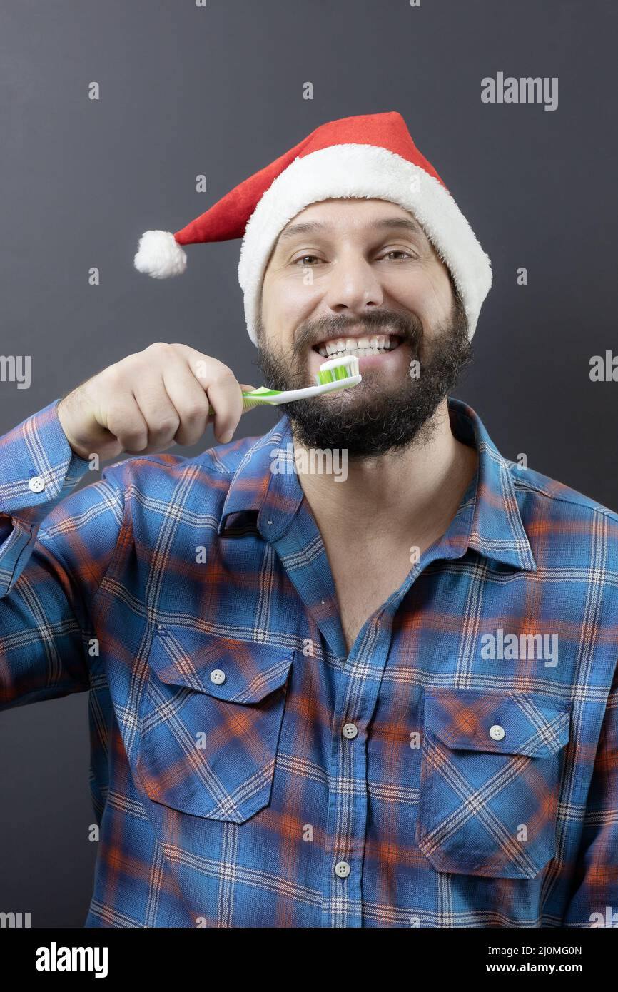 Ein bärtiger Mann in einem Weihnachtsmann-Hut mit einem breiten, weiß gezahnten Lächeln, der sich die Zähne putzt. Vorbereitung auf den Urlaub. Gefälschter weihnachtsmann Stockfoto