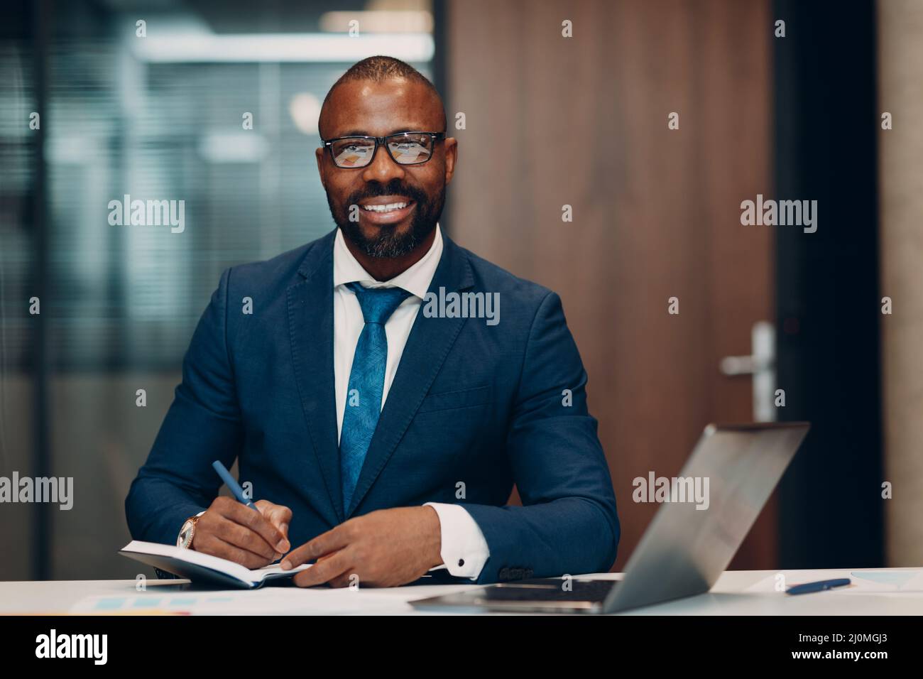 Ein afroamerikanischer Geschäftsmann mit Portrait-Lächeln in blauem Anzug sitzt mit einem Notebook mit Stift und Laptop am Tisch, um sich im Büro zu treffen. Stockfoto