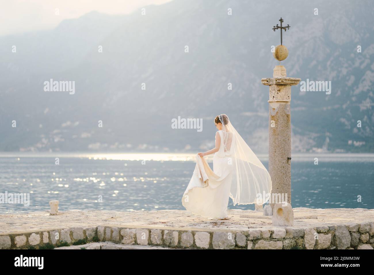 Die Braut in einem langen weißen Kleid und einem im Wind flatternden Schleier wandert entlang der Insel Gospa od Skrpela Stockfoto