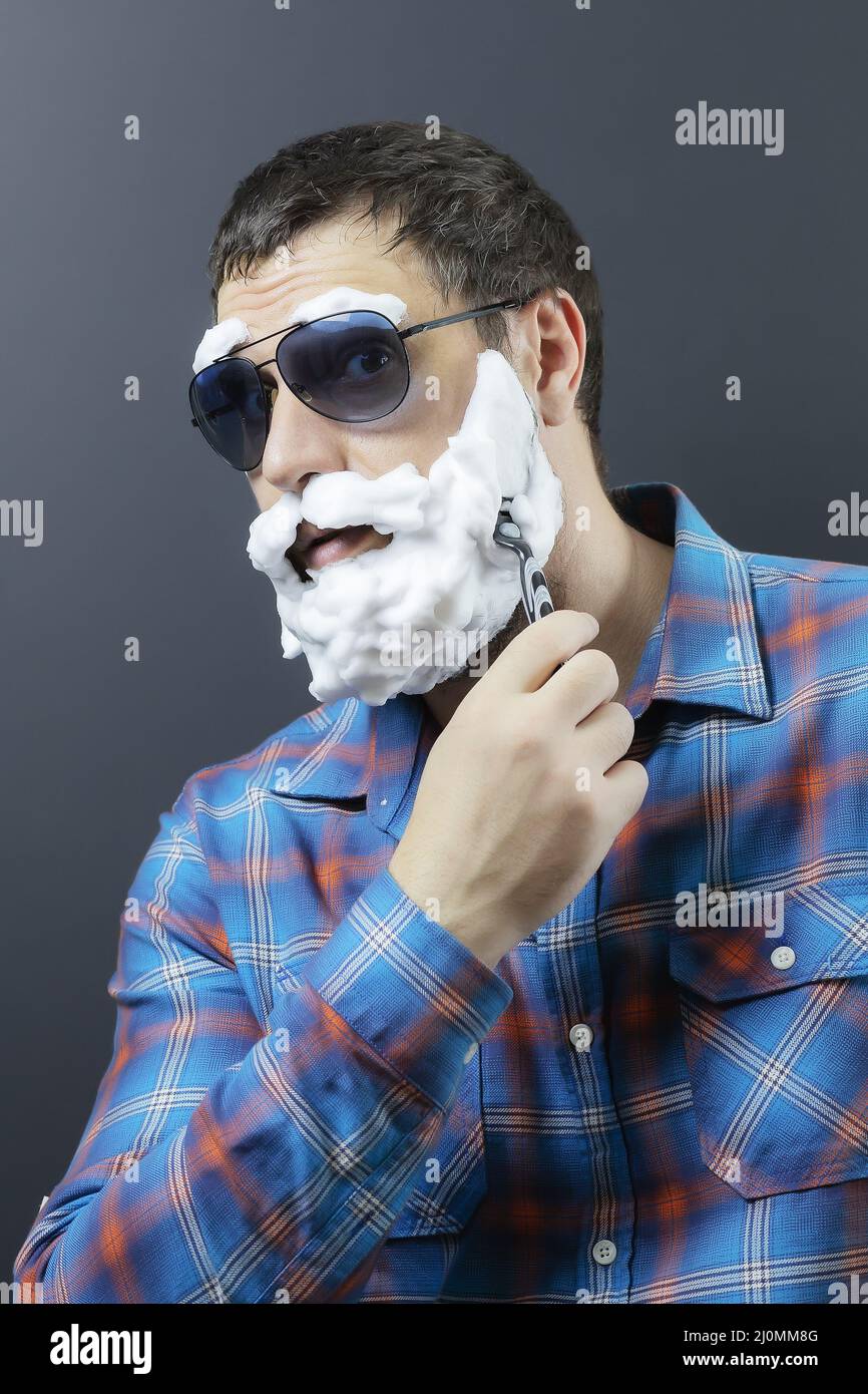 Ein Mann mit einem überraschenden Blick in Sonnenbrille mit Bart und Augenbrauen aus Schaumstoff rasiert mit einem Rasiermesser Stockfoto