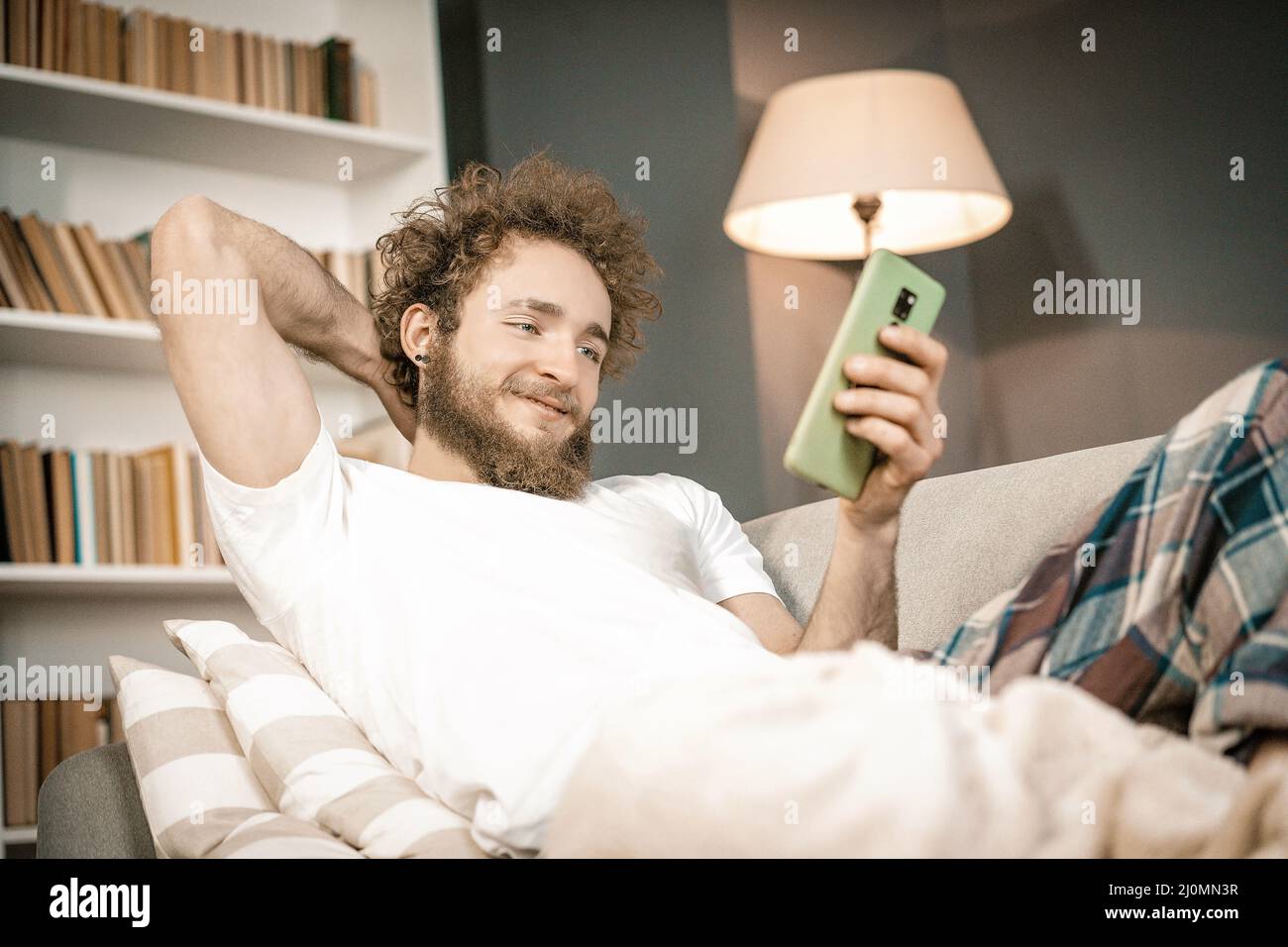 Lockiger Mann, der auf der Couch liegt, auf den Telefonbildschirm schaut und in den sozialen Medien nachschaut. Romantischer Mann auf der Suche nach einem Girlfrie Stockfoto