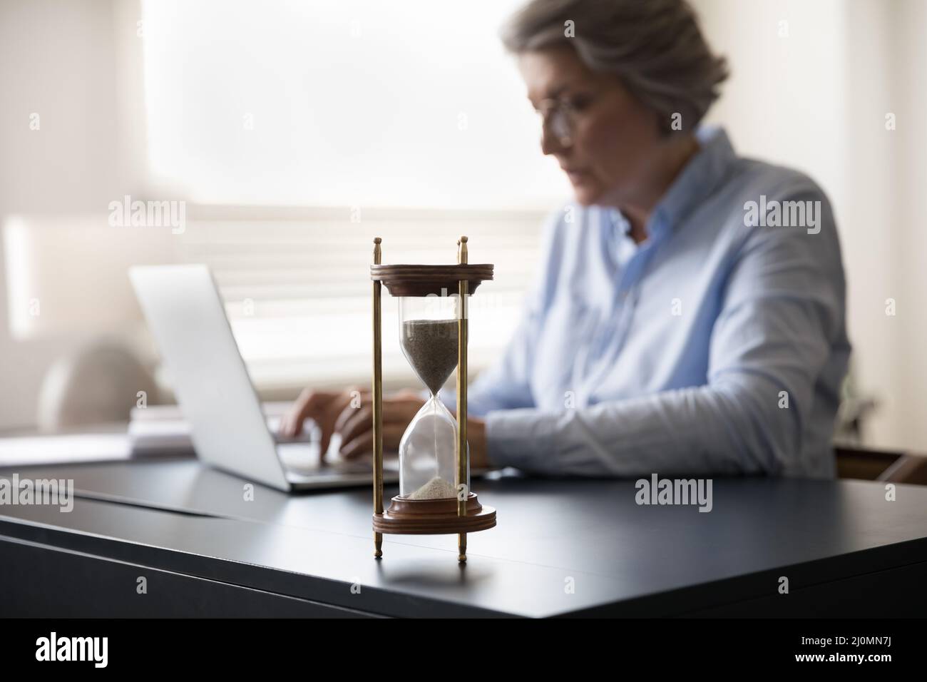 Reife Geschäftsfrau arbeitet am Laptop, Sanduhr auf dem Tisch, Vorderansicht Stockfoto