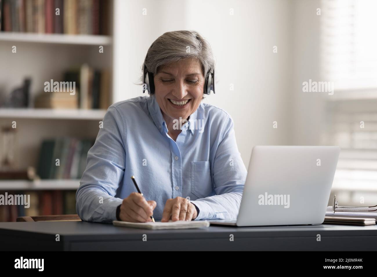 Eine ältere Frau sitzt am Schreibtisch und hört den Ton über Kopfhörer Stockfoto