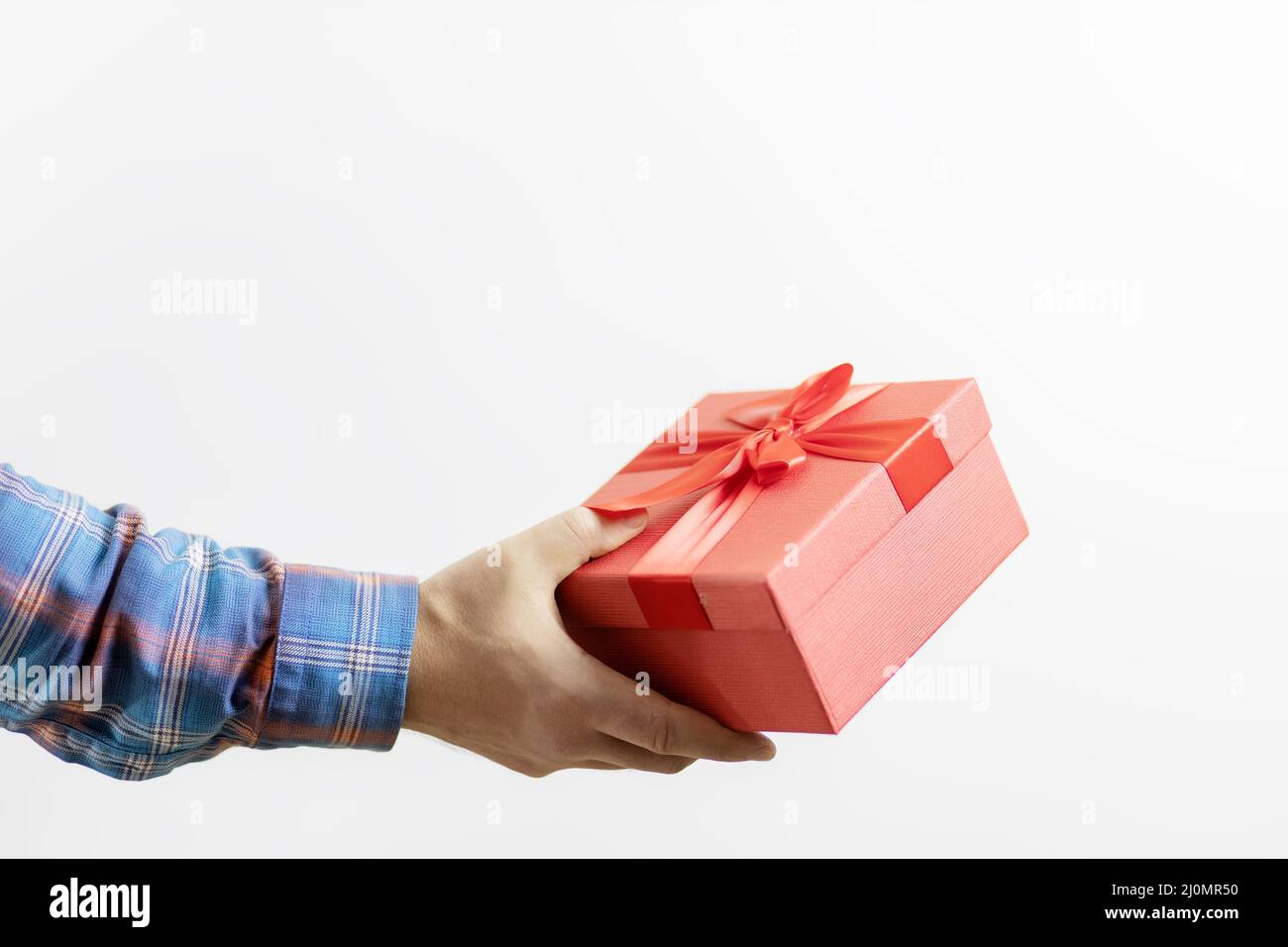 Eine Männerhand in einem karierten Ärmel hält eine rote Schachtel mit einem Geschenk auf weißem Hintergrund. Geschenk für eine Feier Stockfoto