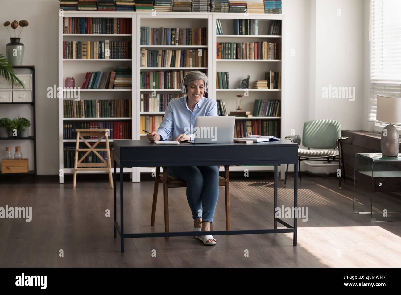 Eine ältere Frau sitzt am Schreibtisch mit einem Laptop, der E-Learns macht, und schreibt Informationen Stockfoto