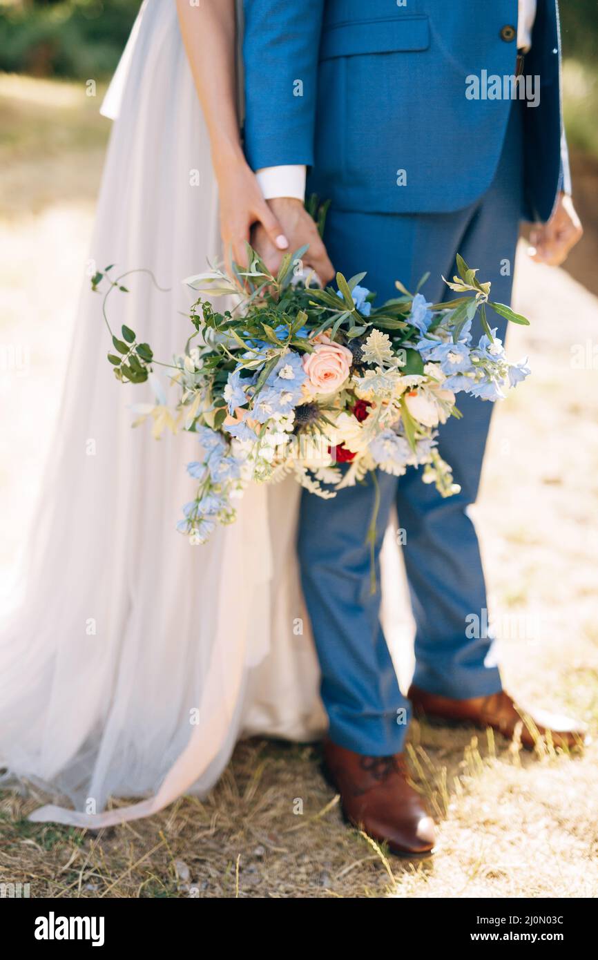 Bräutigam in einem blauen Anzug hält einen Blumenstrauß in der Hand. Braut in einem weißen Kleid umarmt ihn von hinten. Nahaufnahme Stockfoto