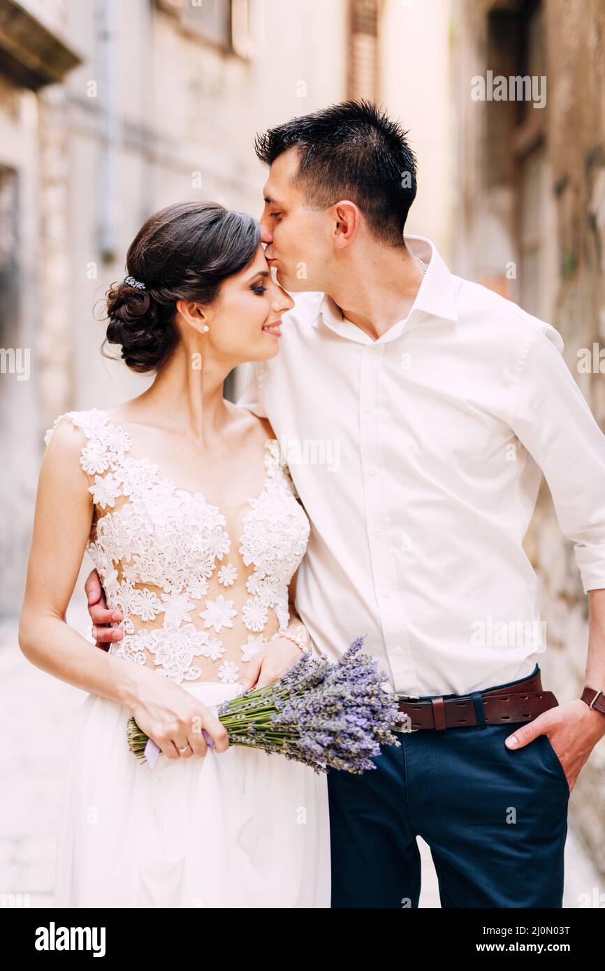 Sibenik, Kroatien - 05.06.17: Bräutigam umarmt und küsst Braut in einer Stirn in einem weißen Spitzenkleid mit einem Strauß Lavendel in ihrem h Stockfoto