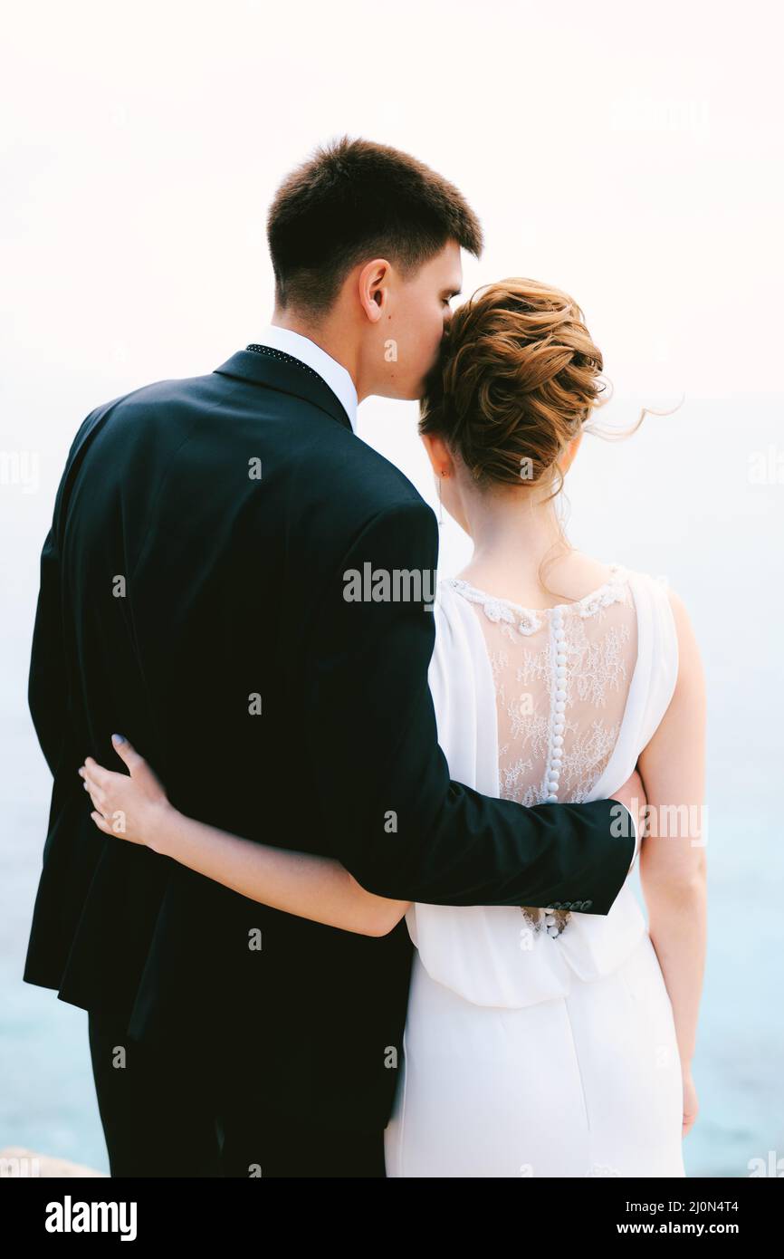Bräutigam in einem schwarzen Anzug umarmt und küsst Braut in einem weißen schönen Spitzenkleid im Tempel. Rückansicht Stockfoto