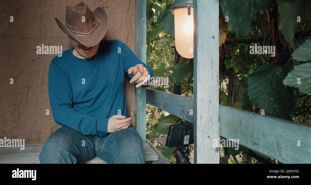 Cowboy-Mann sieht Comedy auf seinem Handy und lacht auf der Veranda des Hauses sitzen Stockfoto