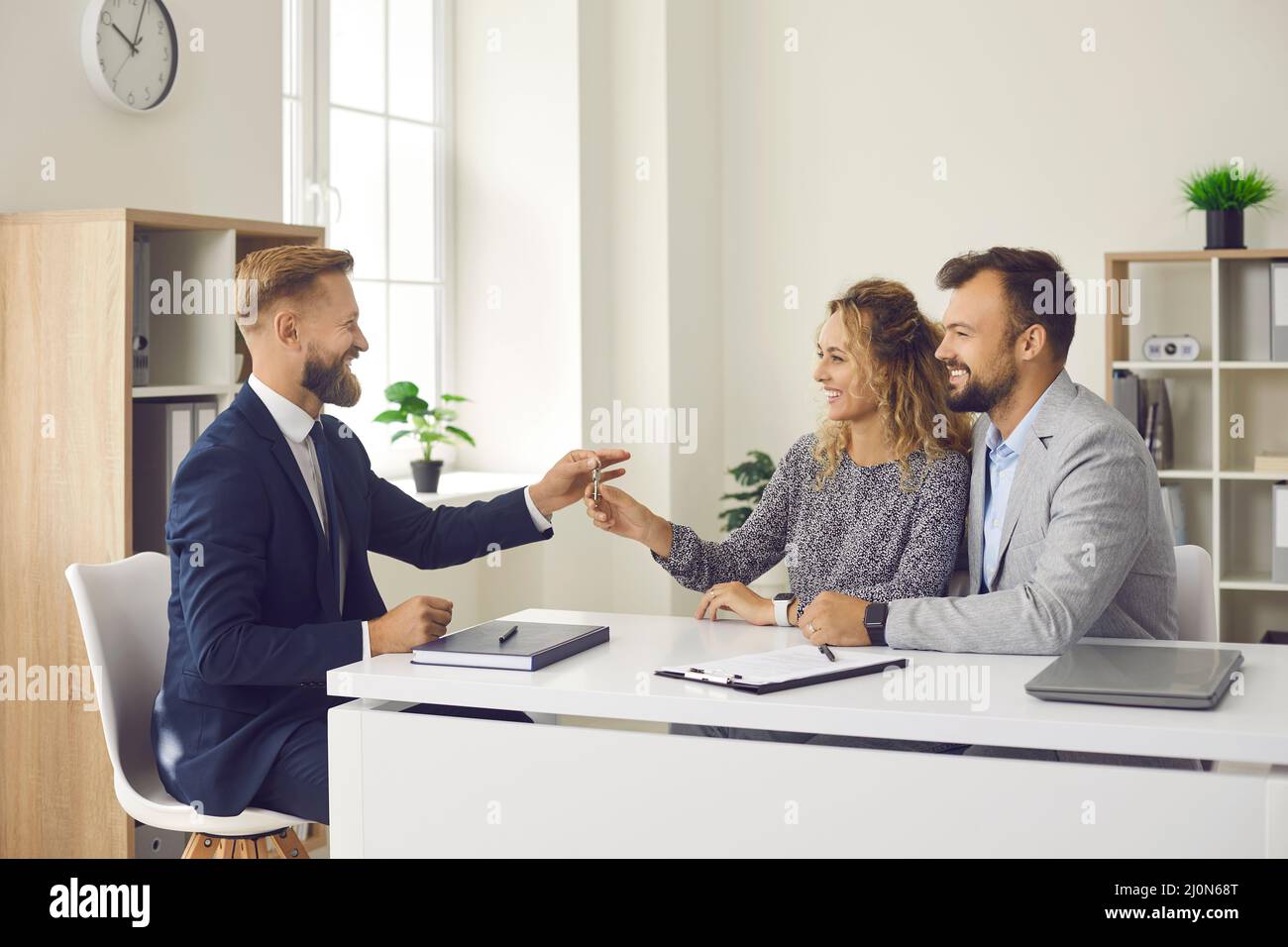 Lächelnder männlicher Immobilienmakler gibt der jungen Familie, die zusammen das erste Haus kaufen, Schlüssel. Stockfoto