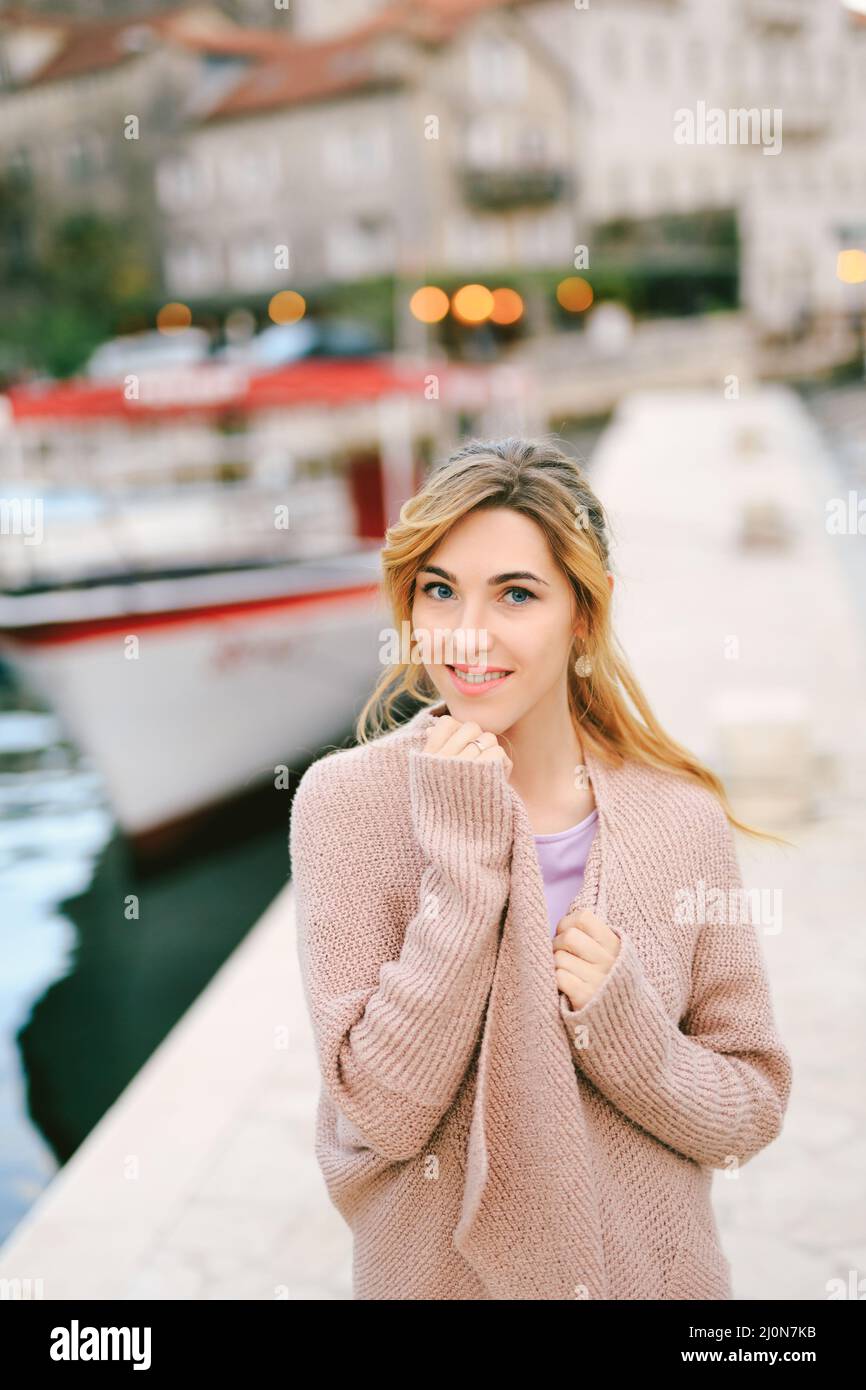 Junge lächelnde Frau, die sich in eine warme Jacke gewickelt hat, während sie vor dem Hintergrund von Booten und Gebäuden auf dem Pier stand Stockfoto