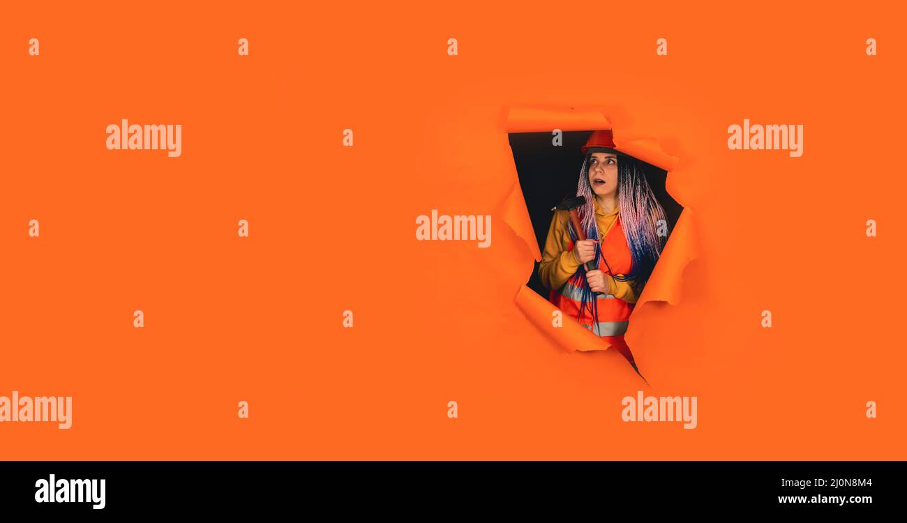 Schockierte Bauarbeiterin in Overalls mit Hammer, der aus einem Loch mit orangefarbenem Hintergrund schaute. Junge Frau, die sich im Schockzustand umsieht Stockfoto
