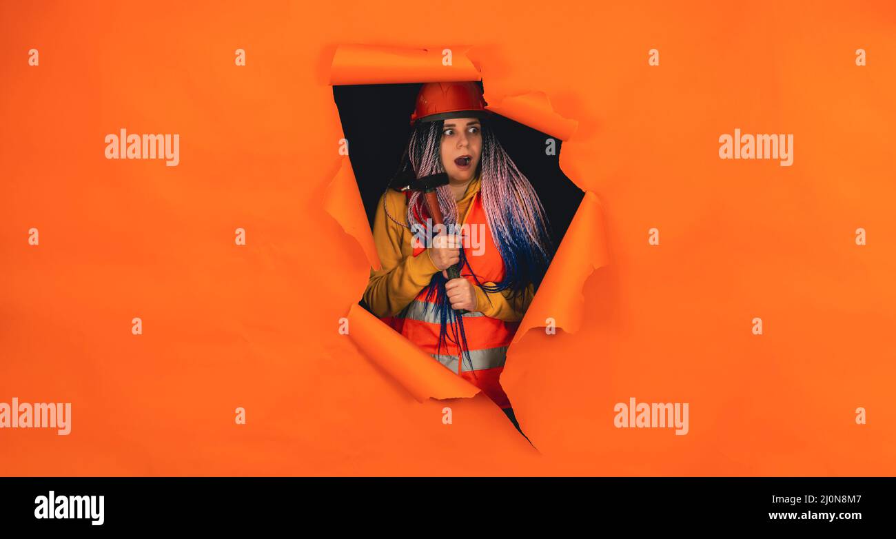 Schockierte Bauarbeiterin in Overalls mit Hammer, der aus einem Loch mit orangefarbenem Hintergrund schaute. Junge Frau, die sich im Schockzustand umsieht Stockfoto
