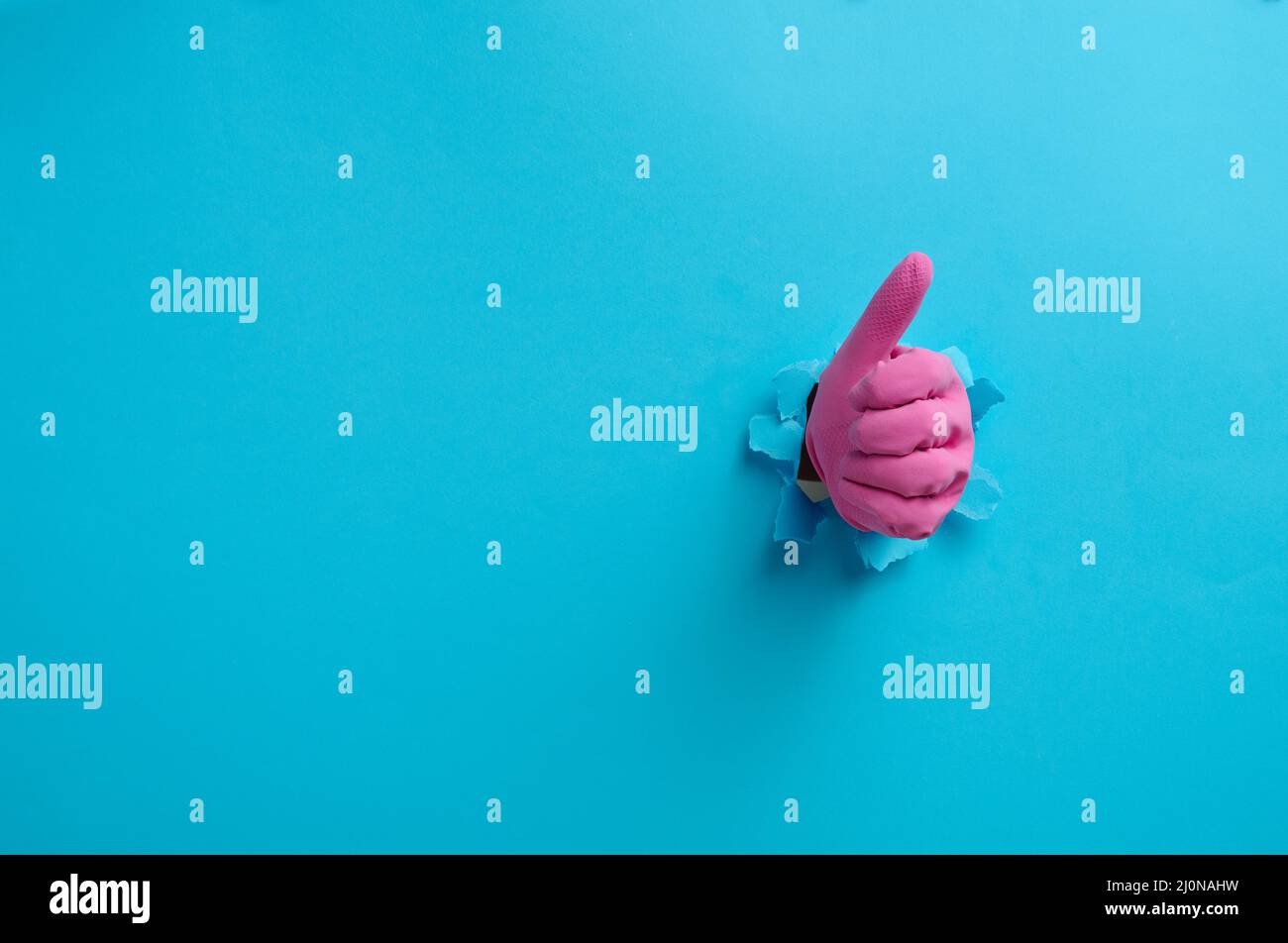 Eine Hand in einem pinken Haushaltsgummihandschuh ragt aus dem zerrissenen Loch und zeigt eine Geste okay, Daumen hoch. Blauer Hintergrund Stockfoto