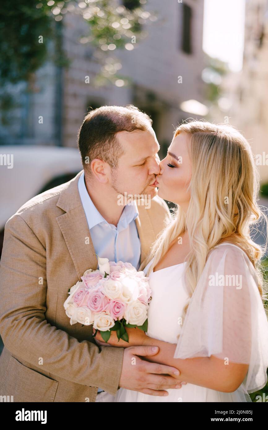 Die Braut mit Blumenstrauß und Bräutigam umarmen und küssen sich in einer gemütlichen Straße der Altstadt aus nächster Nähe Stockfoto
