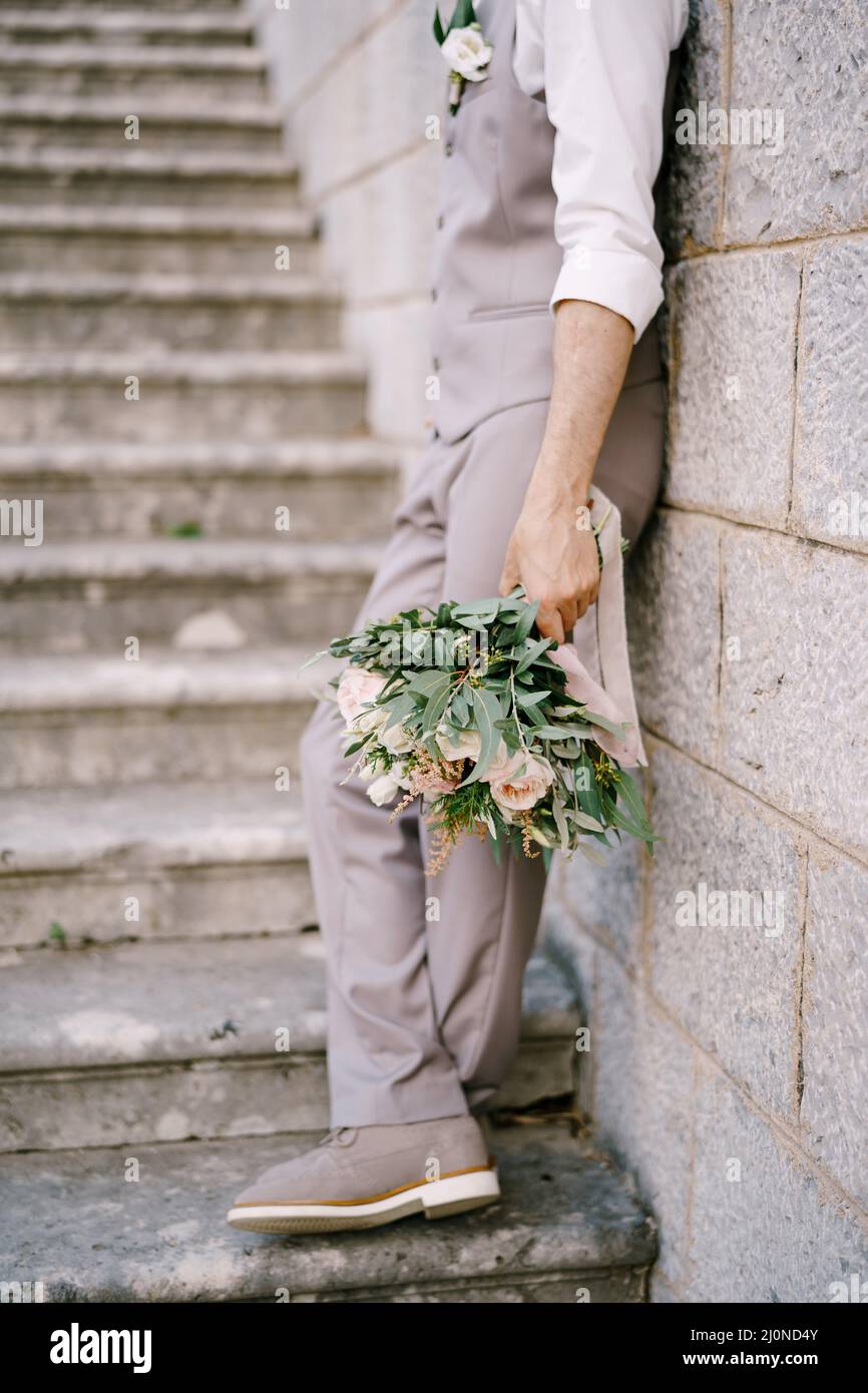 Bräutigam mit einem Strauß steht auf den Steinstufen, lehnt an der Wand Stockfoto