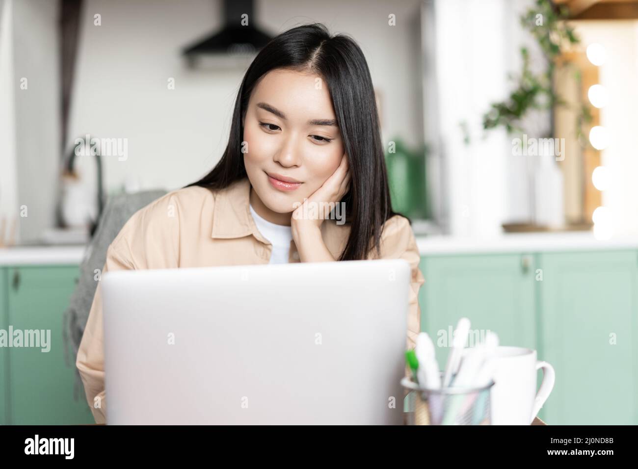 Lächelnde asiatische Frau, die sich entspannt beim Webinar ansieht, während sie zu Hause auf den Laptop schaut Stockfoto