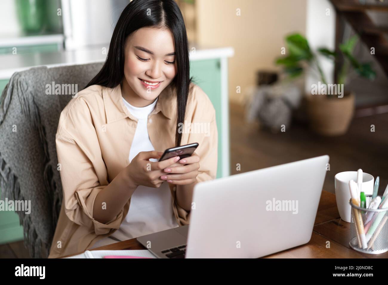 Asiatische Studentin mit Blick auf Mobiltelefon, zu Hause mit Laptop sitzen, Online-Kurs auf Laptop registrieren, Remote arbeiten Stockfoto