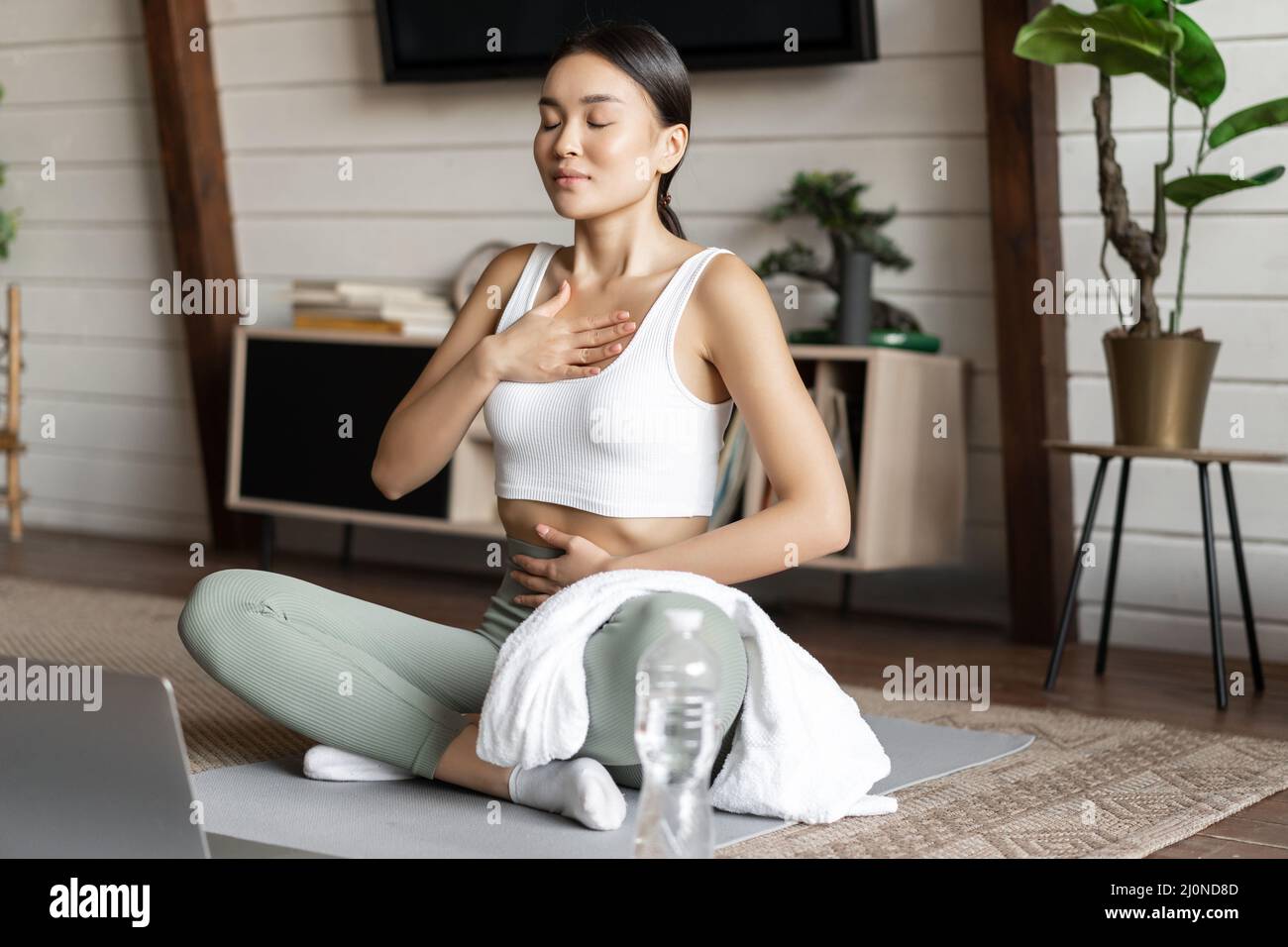 Meditation und Online-Klassen Konzept. Junge Frau, die zu Hause ferntrainiert, im Wohnzimmer auf einer Gummimatte sitzt und Lapto verwendet Stockfoto