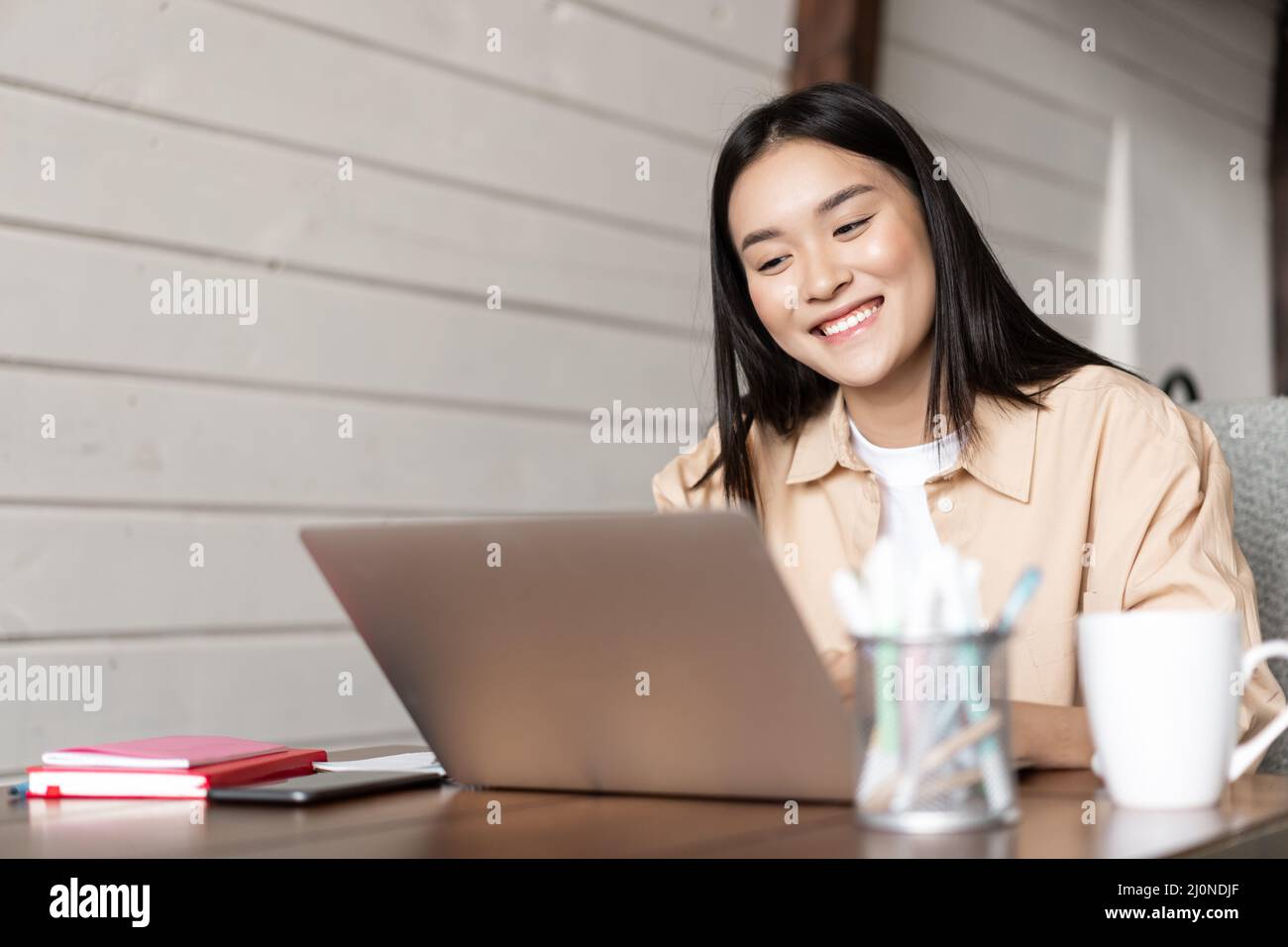 Lächelndes asiatisches Mädchen schaut auf den Laptop-Bildschirm, arbeitet von zu Hause aus, hört sich Webinare an oder nimmt an Online-Vorlesungen Teil, an Universitätskursen auf rem Stockfoto