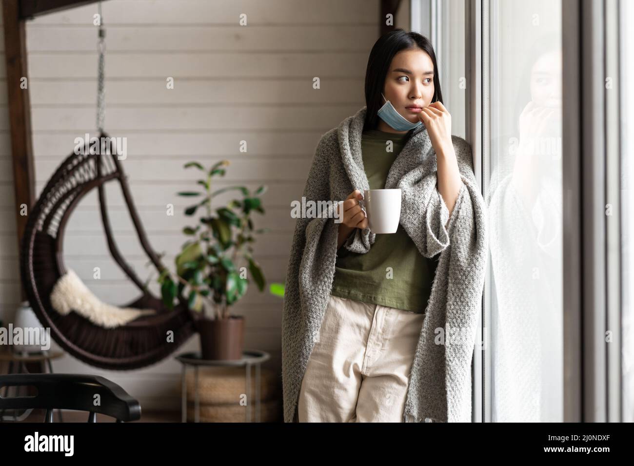 Junge asiatische Frau, die Tee trinkt und zu Hause aus dem Fenster auf Quarantäne schaut, weil sie an einer Covid-19-Pandemie erkrankt ist Stockfoto