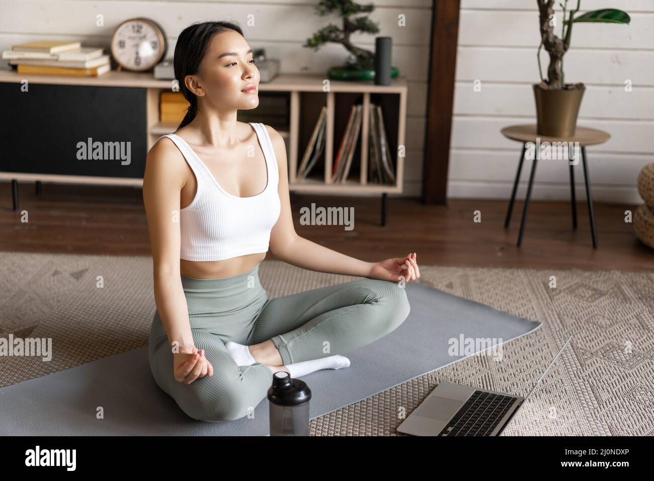 Junge asiatische Frau, die Yoga mit Laptop macht, Meditationslehrer auf der Anwendung oder Website hört, Meditationskurs macht Stockfoto