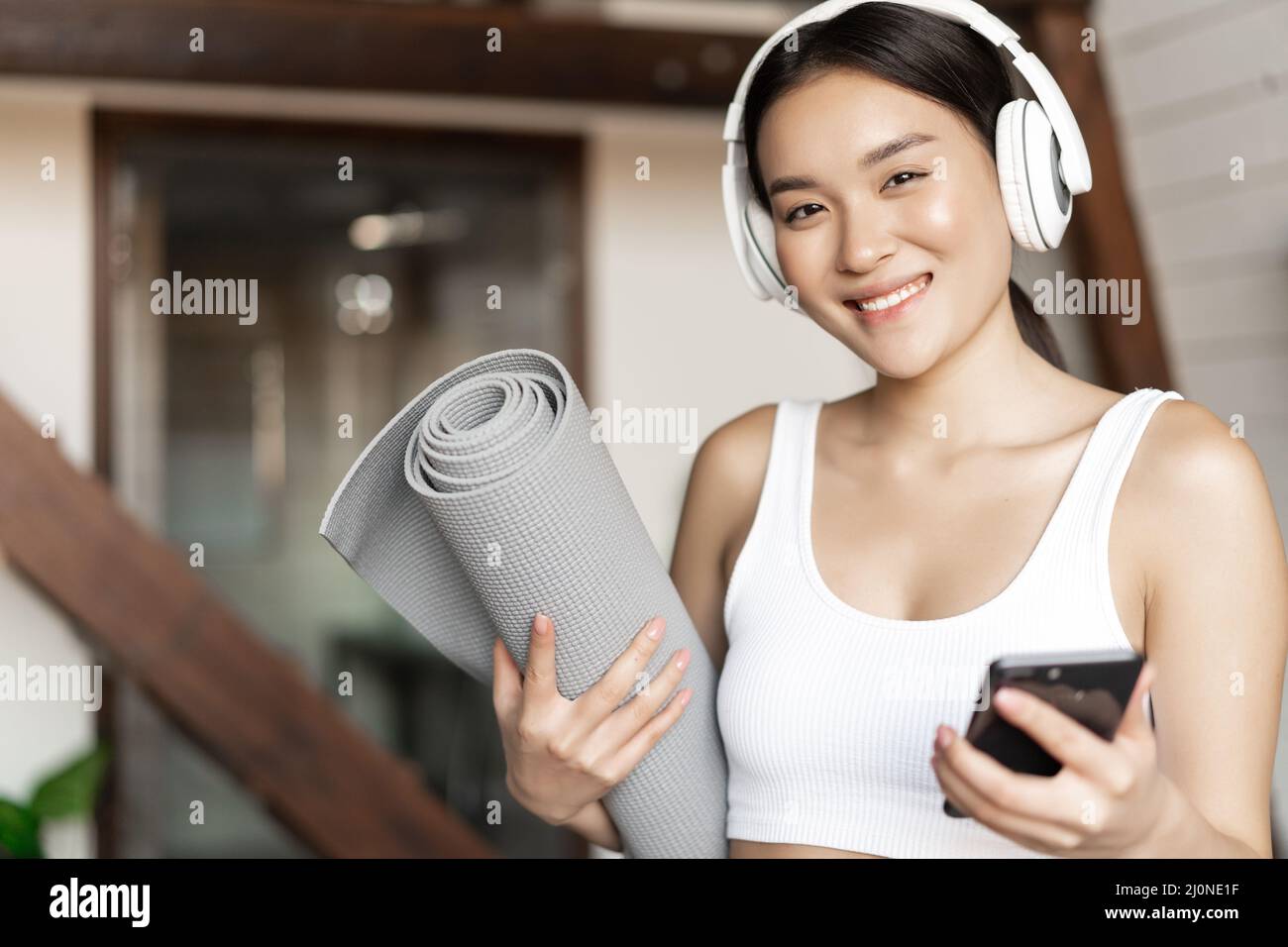 Lächelndes asiatisches Mädchen in Kopfhörern, Musik zu Hause hören und mit einer Bodenmatte trainieren, Mobiltelefon halten, Fitnessübungen machen Stockfoto