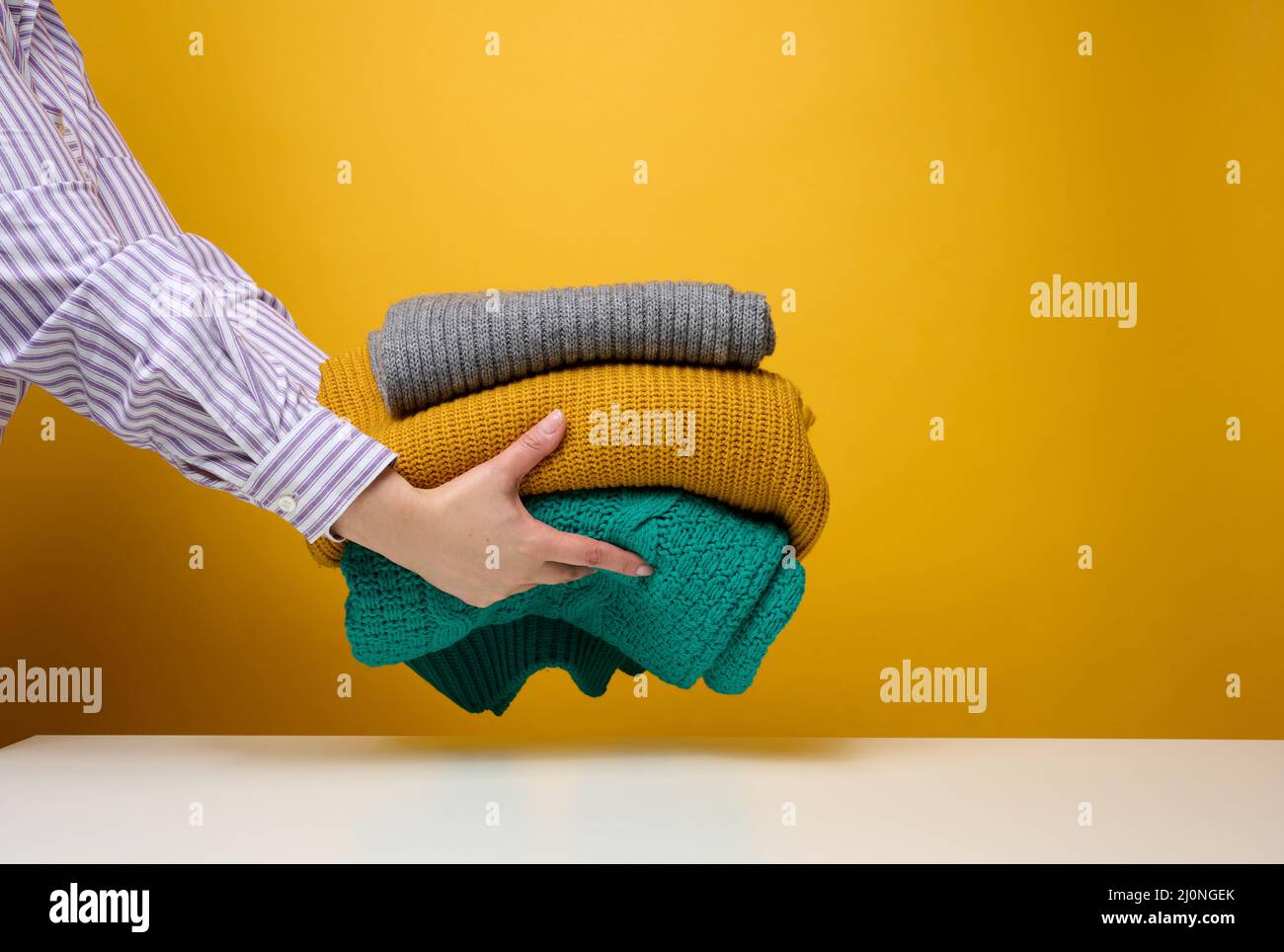 Frau mit einem Stapel gestrickter, gefaltetem Pullover auf gelbem Hintergrund. Wäsche waschen, Wäsche putzen Stockfoto