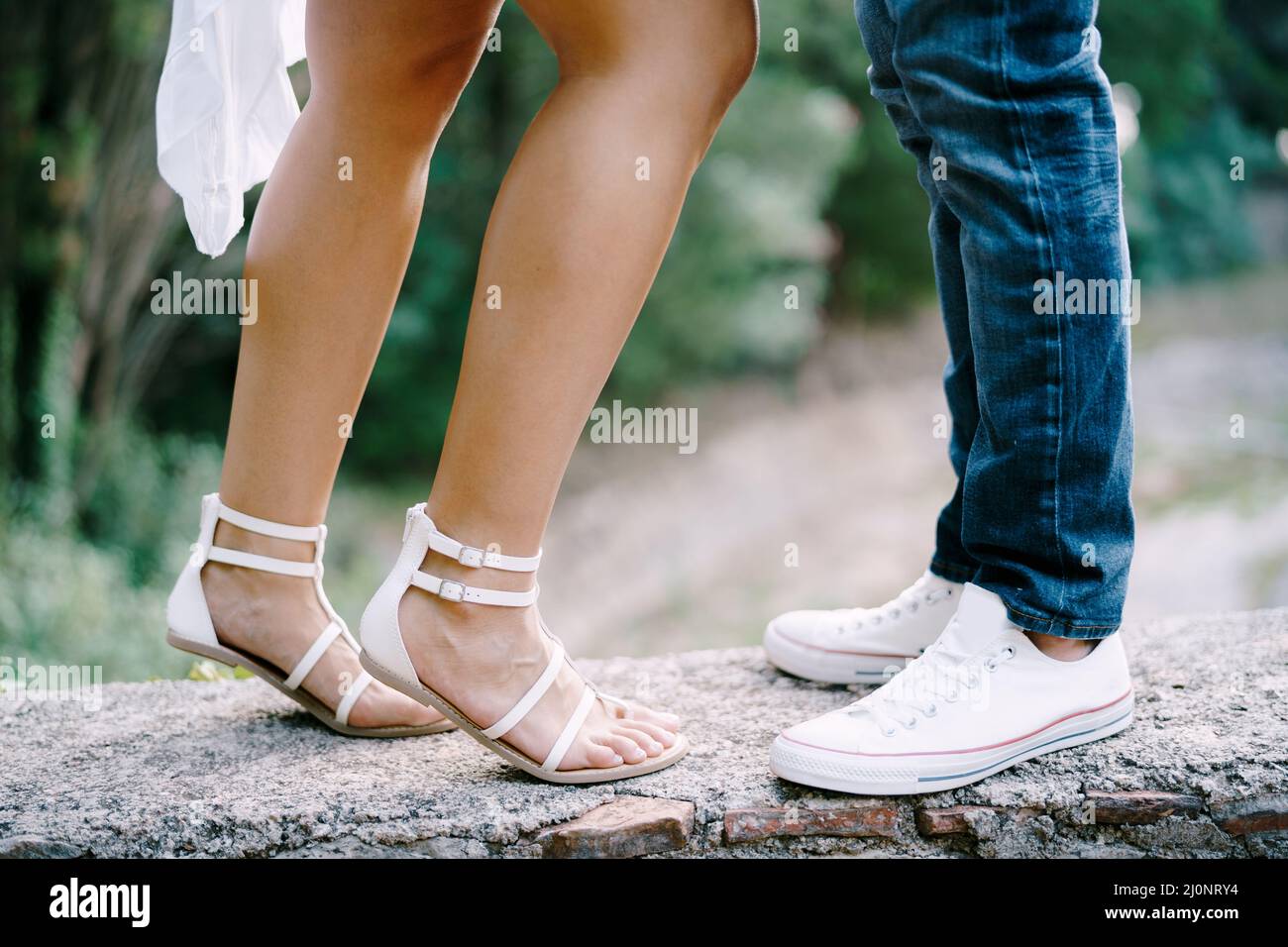 Weibliche Füße in Sandalen neben männlichen Füßen in Sneakers stehen an einer Steingrenze im Park Stockfoto