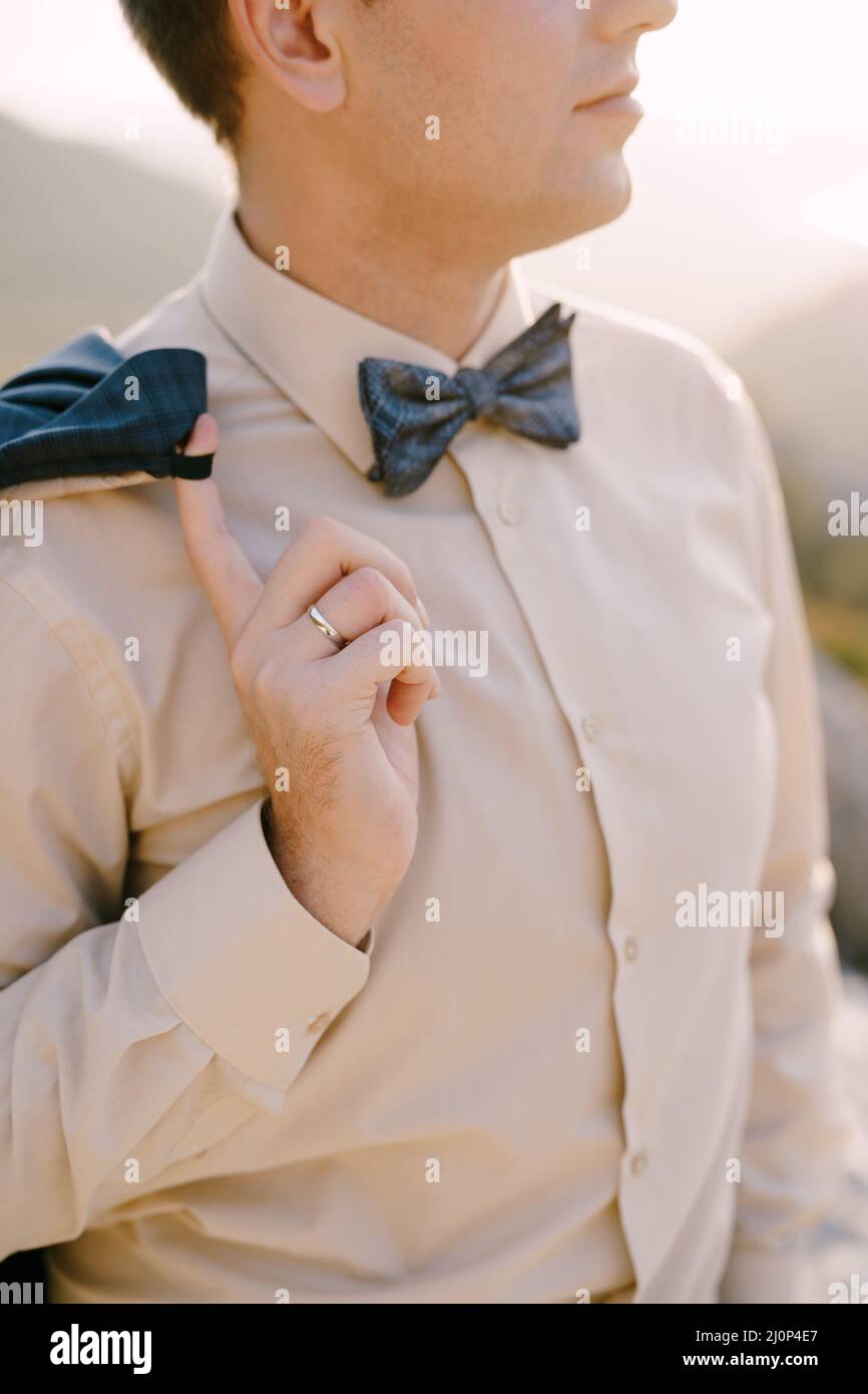 Bräutigam in einem beigen Hemd und Fliege hält seine Jacke mit dem Finger auf der Schulter. Nahaufnahme Stockfoto