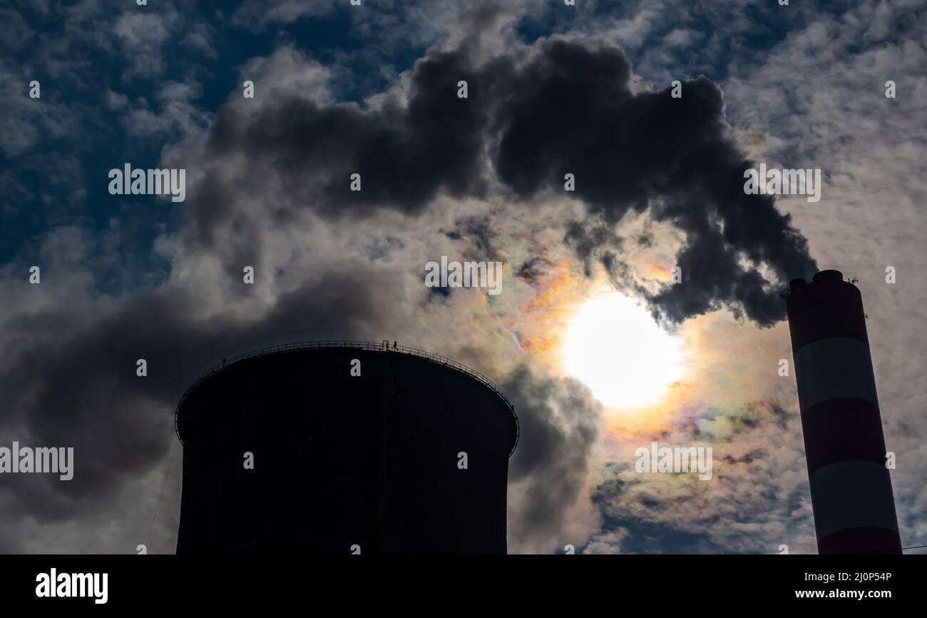 Eine Nahaufnahme des Rauchs und des Dampfes, der aus den Schornsteinen eines Kohlekraftwerks austritt. Das Foto wurde an einem sonnigen Tag gegen das Licht aufgenommen, das Stockfoto