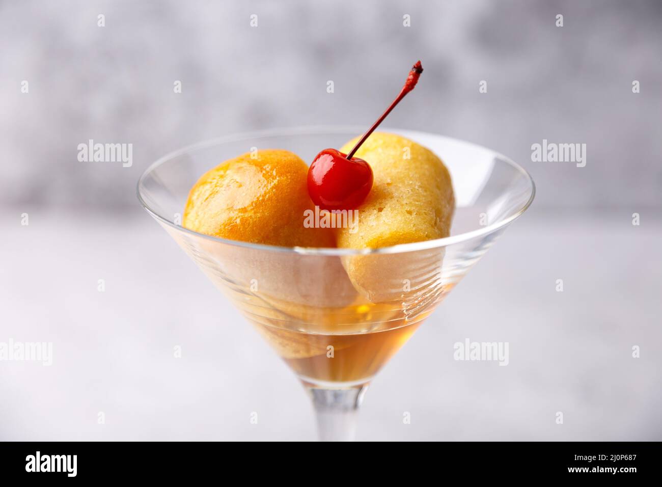 Neapolitanischer Rum baba (oder baba au Rum) in einem martini-Glas mit einer Cocktailkirsche auf grauem Hintergrund. Kleine Hefekuchen in Rumsirup getränkt. Traditiona Stockfoto