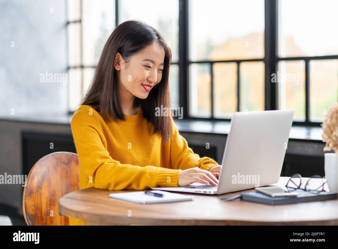 Asiatische Frau, die zu Hause oder im Café am Laptop arbeitet. Junge Dame im leuchtend gelben Pullover Stockfoto