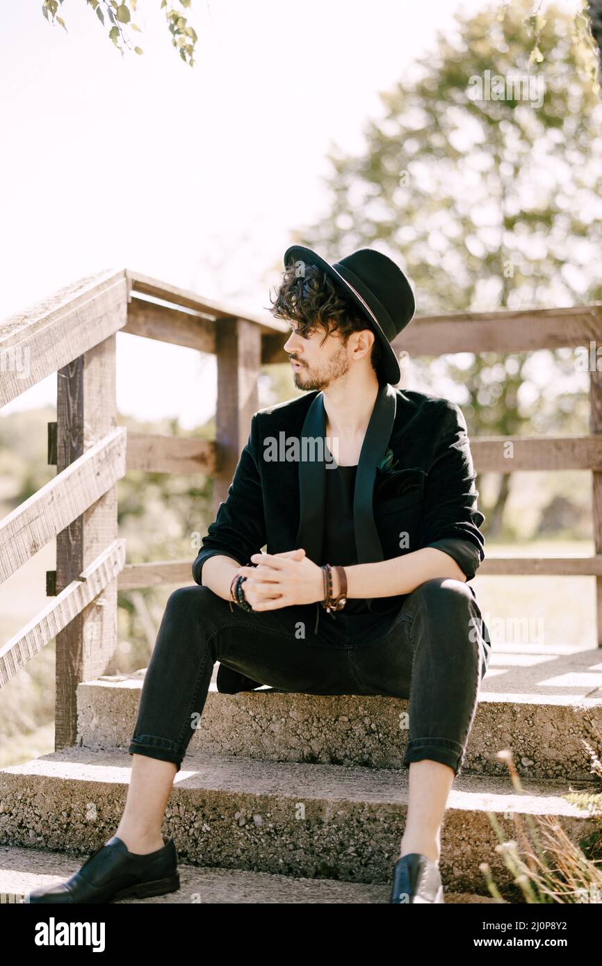 Junger Mann in schwarzem Hut sitzt auf Steintreppen mit Holzgeländer Stockfoto