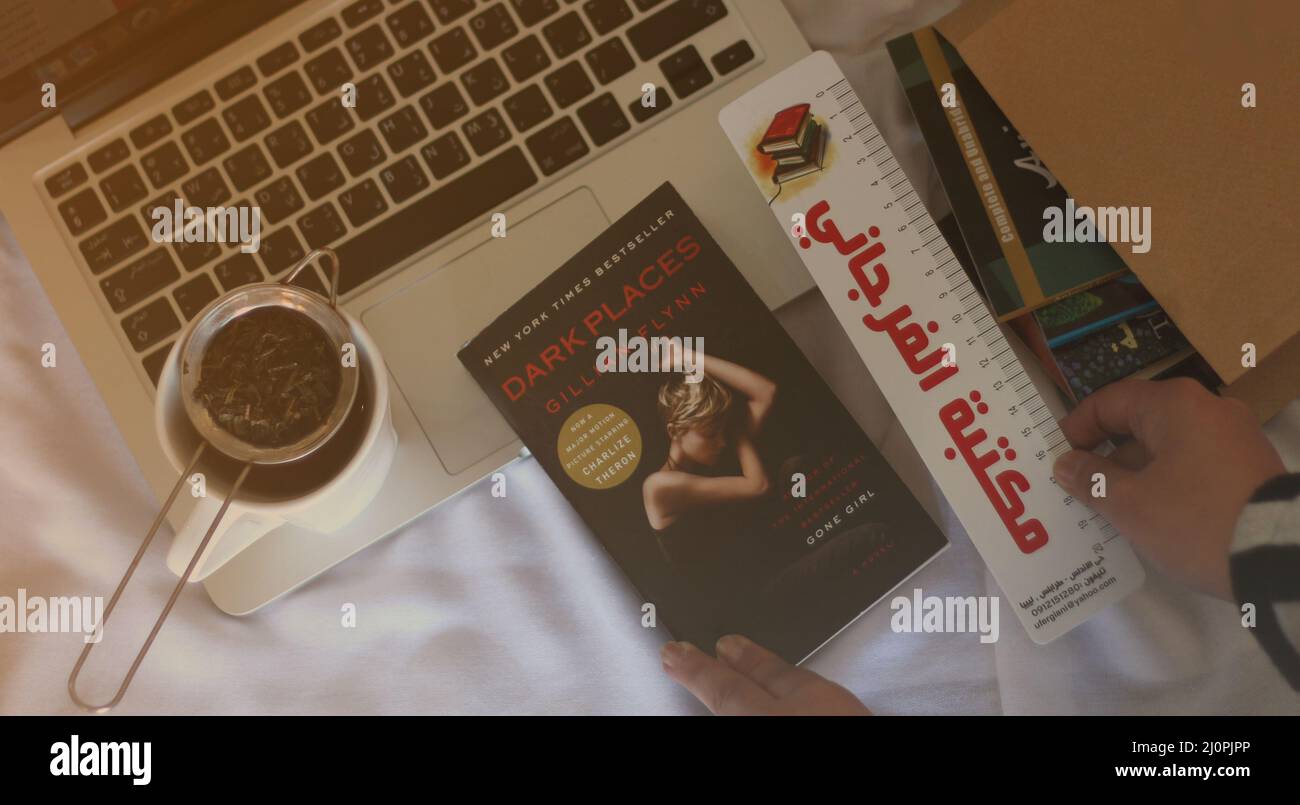 Ein Mac mit einer Tasse Tee, einem Roman und zwei Händen, Sie setzen einen Buchtrenner in den Roman, und auf der Seite ist eine Papiertüte mit einem Satz von Büchern darauf Stockfoto