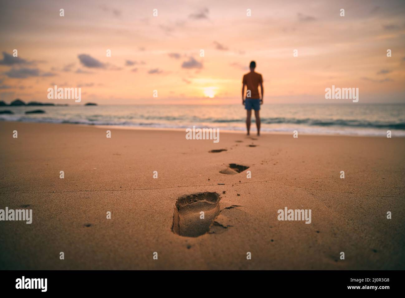 Fußabdrücke im Sand gegen die Silhouette der Person. Einsamer Mann, der bei goldenem Sonnenuntergang am Strand entlang zum Meer geht. Stockfoto