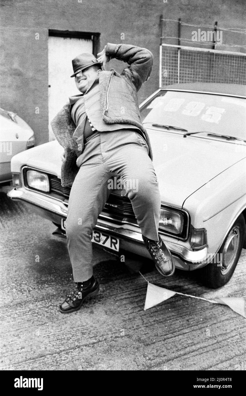 Alexei Sayle, der Komiker aus der Liverpooler Welt, der in der BBC-Fernsehkomödie The Young Ones spielt, besucht einen Autohändler aus zweiter Hand in London. Hier ist er in humorvoller Pose neben einer alten Ford Cortina Limousine abgebildet. 28.. Februar 1984. Stockfoto