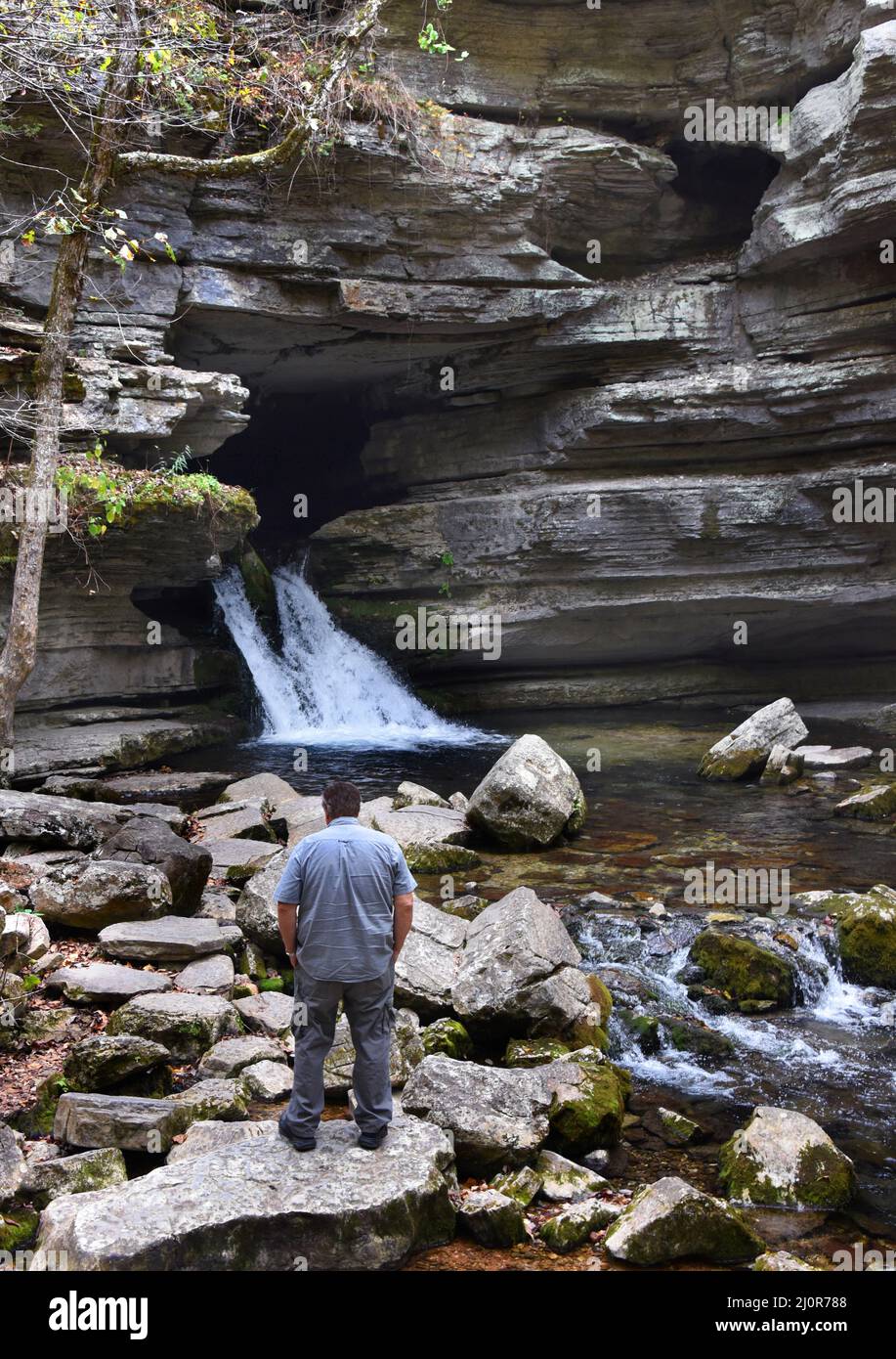 Der Besucher der Blanchard Springs Caverns steht auf einem großen Felsen und bewundert den Wasserfall. Er hat eine graue Hose und ein graues Hemd und hat Hände in den Taschen. Stockfoto
