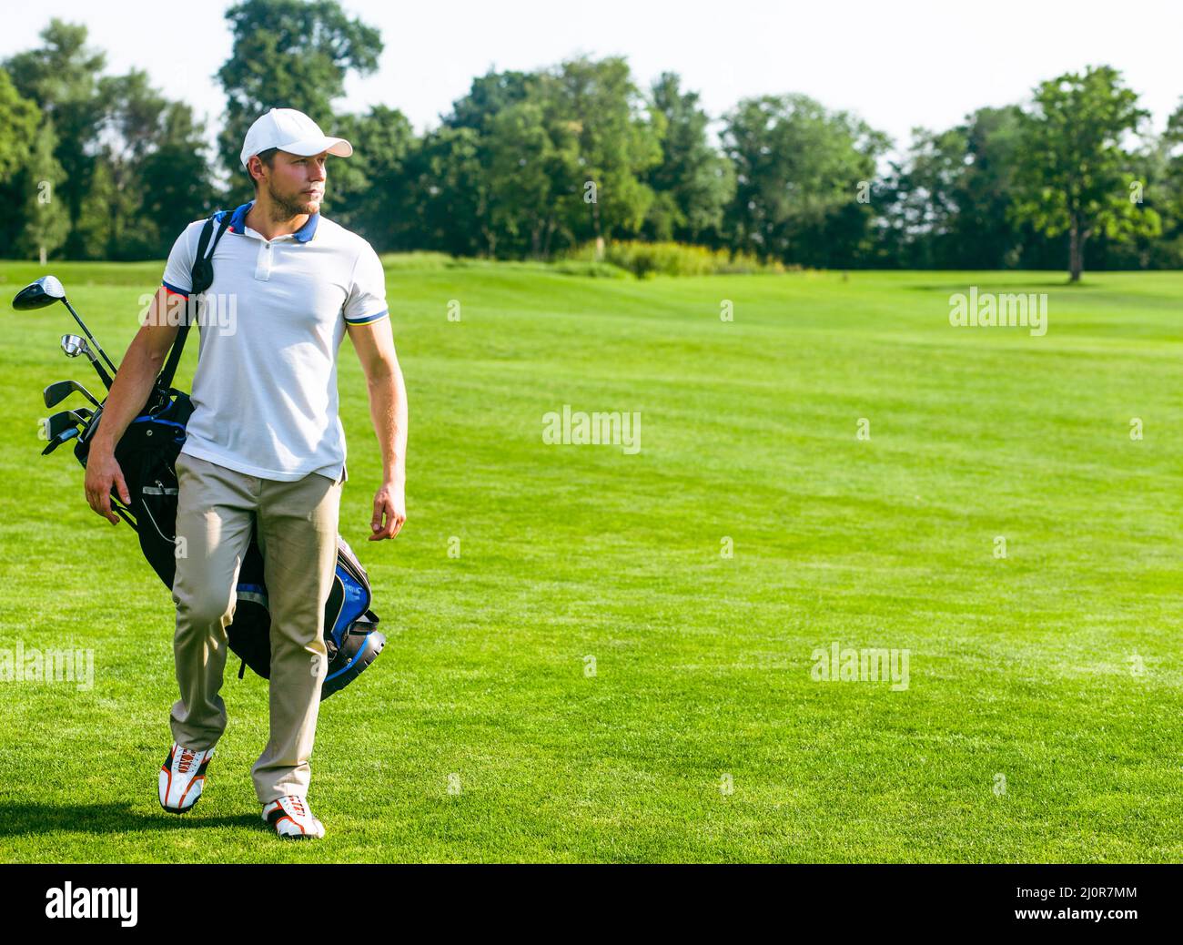 Der junge Golfer trägt eine Tasche mit Golfschlägern. Slim man in Weiß T-Shirt, Golfschuhe, White Cap und Beige Hosen geht in die Stockfoto